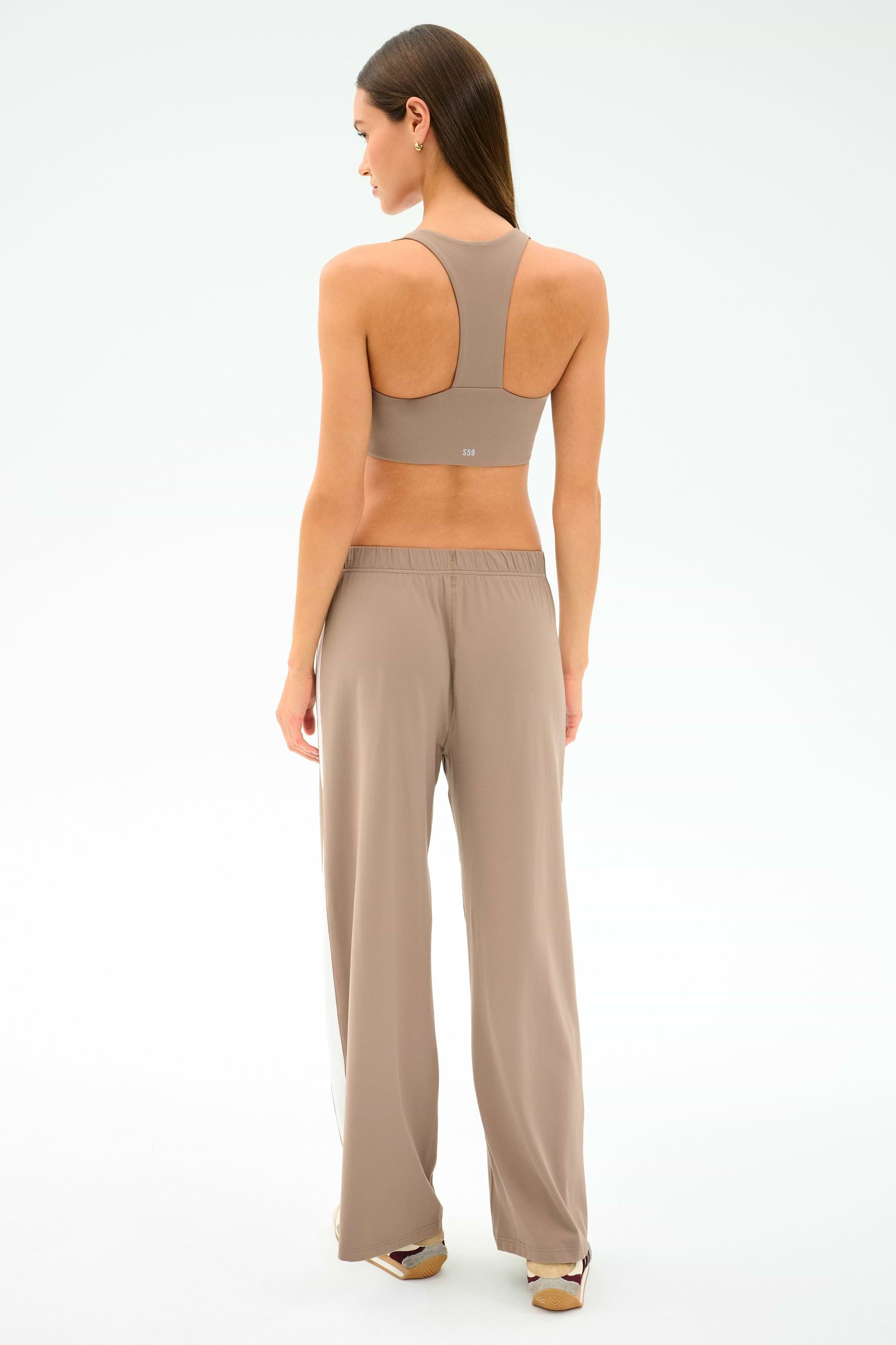 A woman stands facing away, wearing the SPLITS59 Willow Airweight Wide Leg in Latte/White with matching sports bra; her hair is down, shoes visible, against a plain light background.