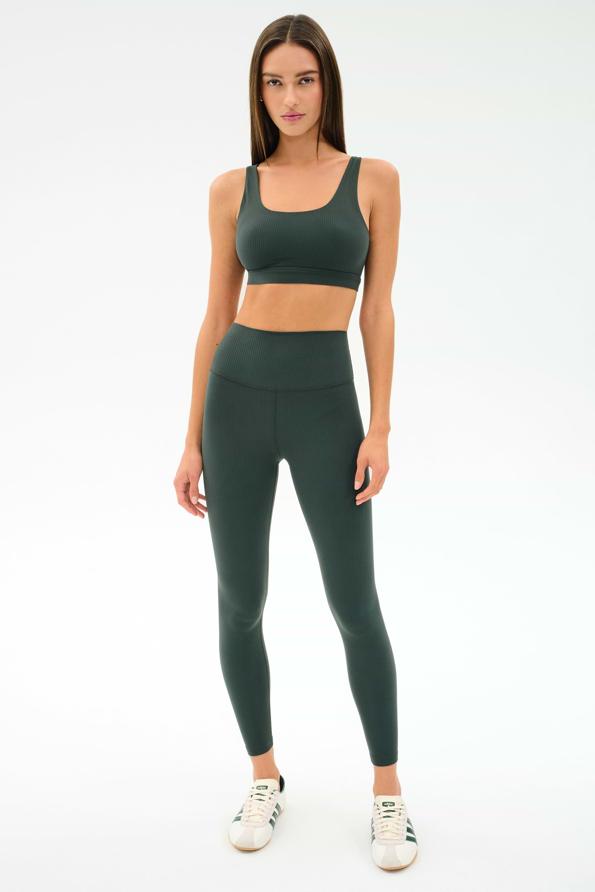 A woman faces forward wearing the SPLITS59 Airweight Rib High Waist 7/8 leggings in Military, paired with a matching sports bra and white sneakers against a plain white background.