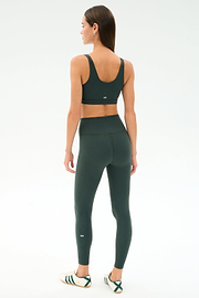 A woman faces away, wearing the SPLITS59 Airweight Rib Bra - Military, matching high-waisted leggings, and white sneakers with green stripes against a plain white background.