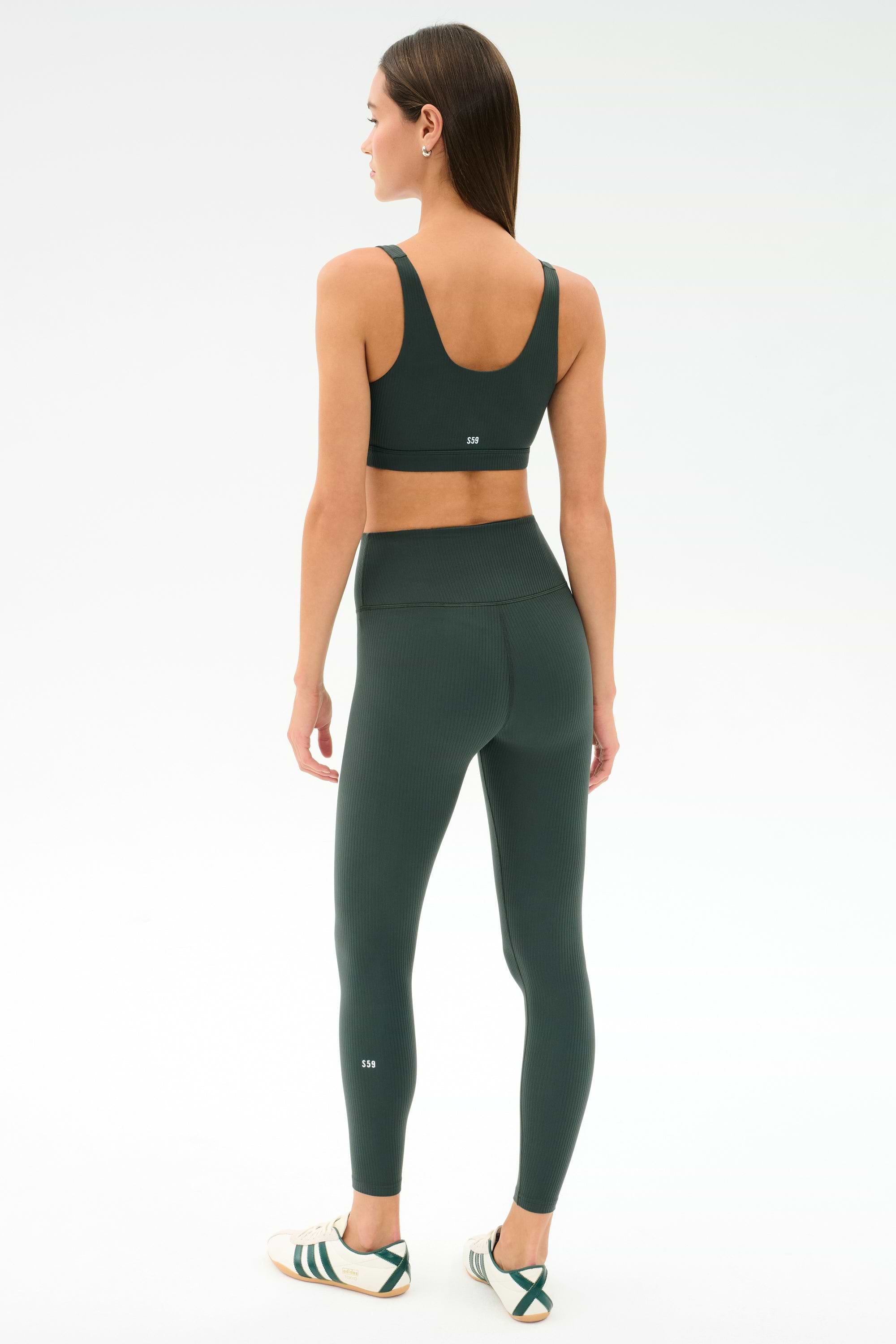 A woman faces away, wearing SPLITS59's Airweight Rib High Waist 7/8 leggings in Military with a matching sports bra and white sneakers, set against a plain white background.
