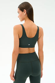 A woman wearing the SPLITS59 Airweight Rib Bra - Military stands facing away from the camera, paired with matching dark green high-waisted leggings made from soft nylon spandex against a plain background.