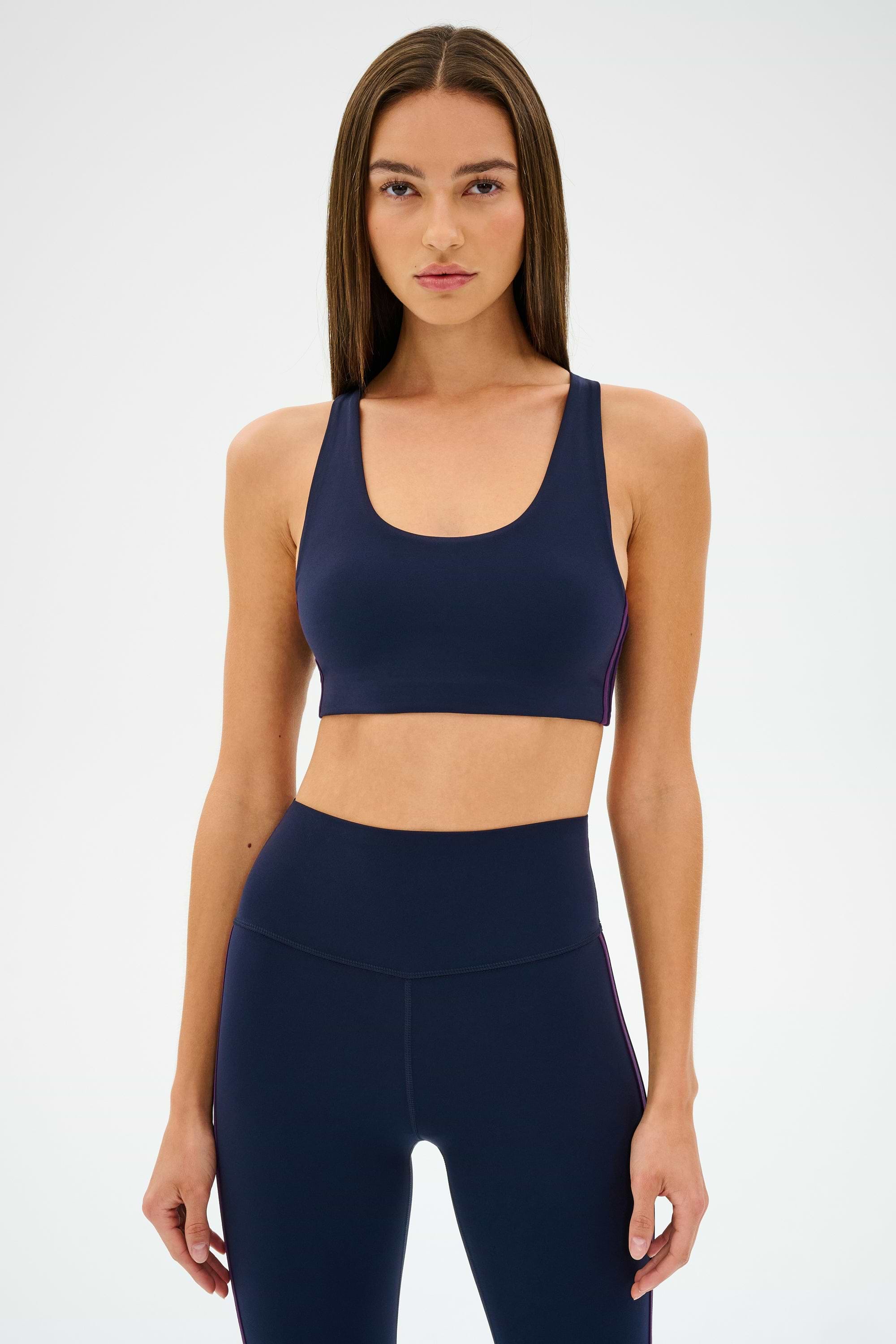 An individual in an Ella Airweight Bra by SPLITS59, in an Indigo/Black Currant color scheme, stands against a plain backdrop, highlighting its sleek racer back design.