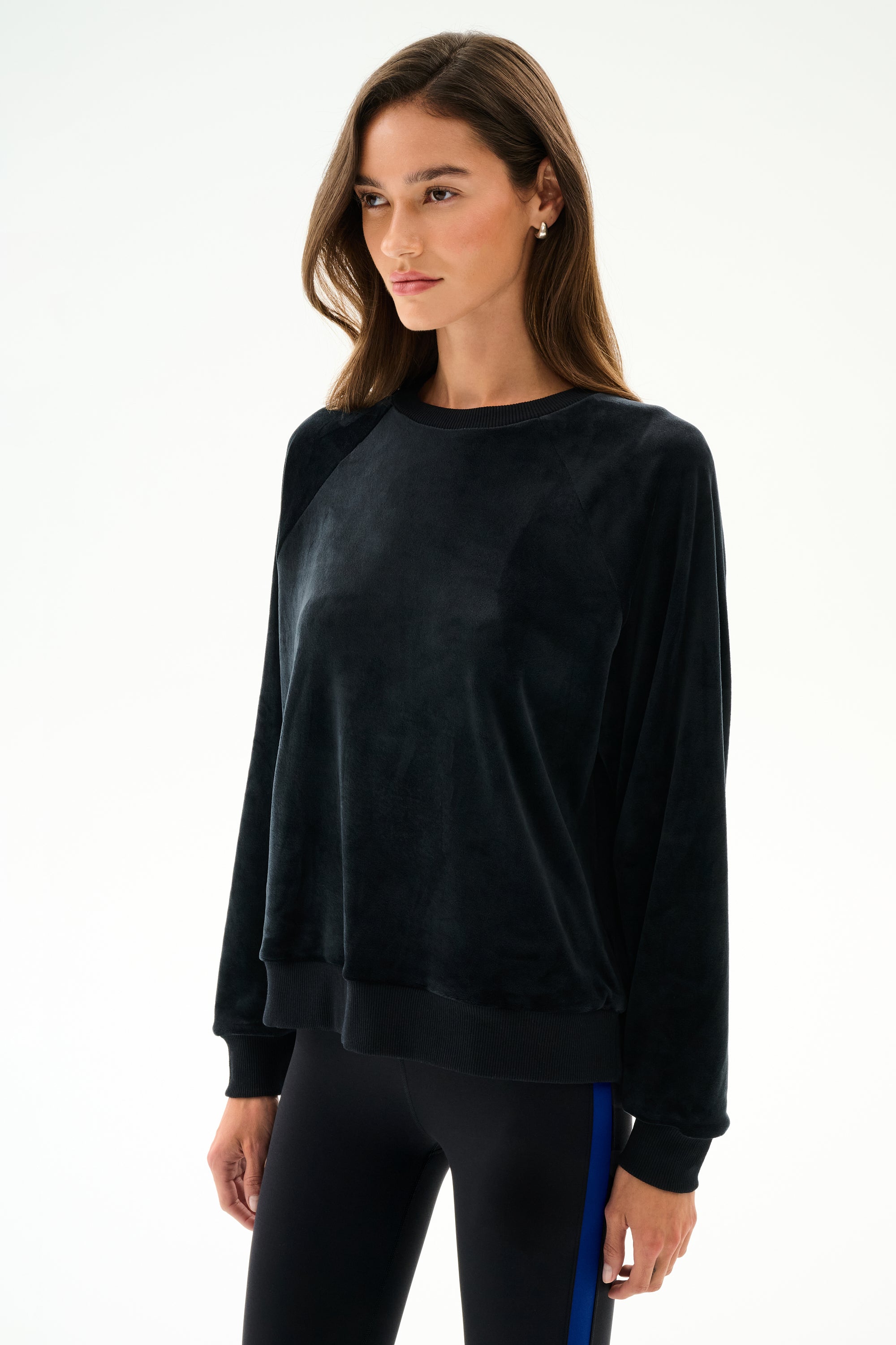 A woman wears the SPLITS59 Joy Velour Crewneck - Black with black leggings featuring a blue stripe, standing against a plain white background.
