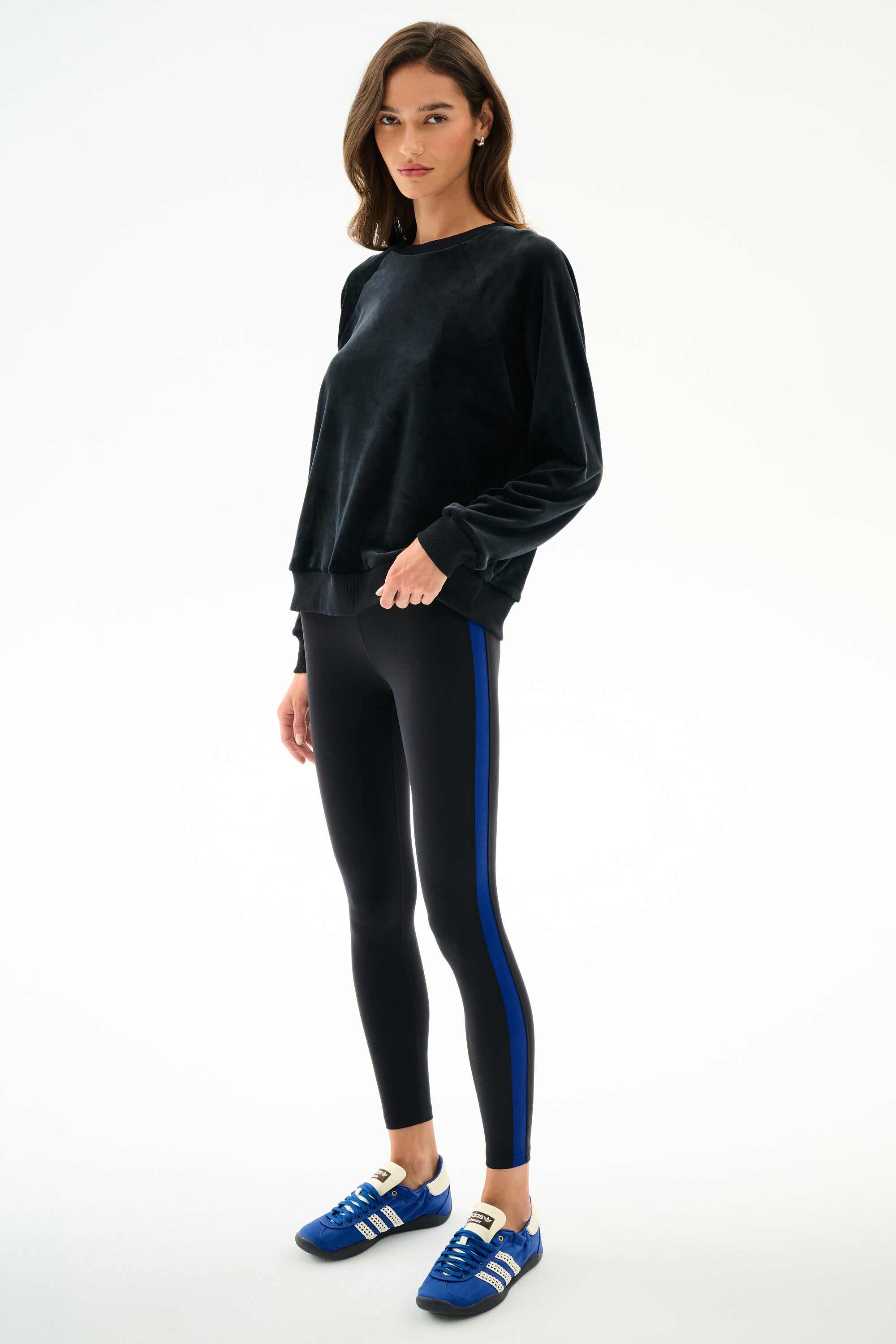 Woman wearing the Joy Velour Crewneck - Black by SPLITS59 and black leggings with a blue stripe, paired with blue Adidas sneakers.