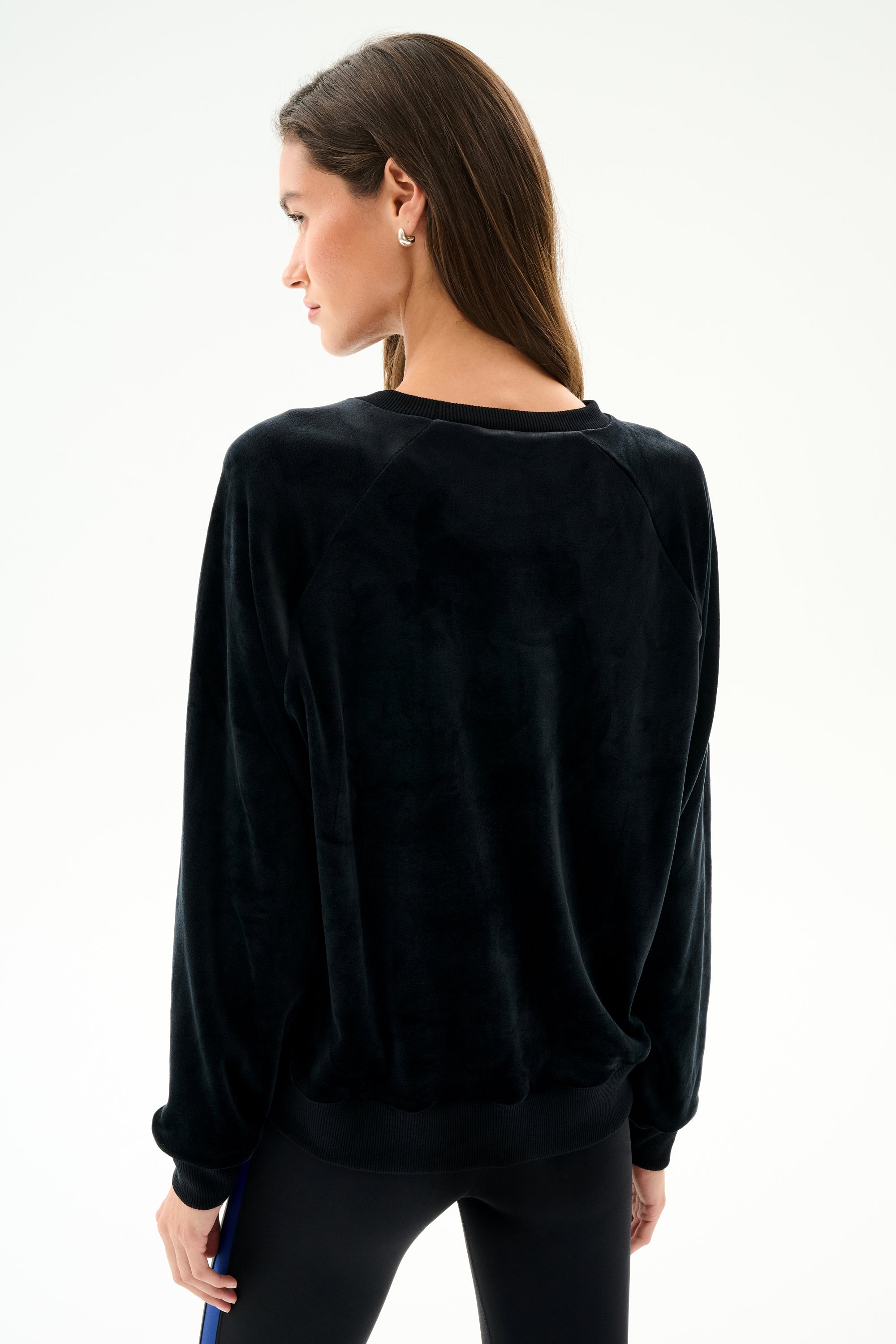 A woman with long brown hair wears the SPLITS59 Joy Velour Crewneck - Black and black polyester spandex leggings, standing against a plain white background, facing away.