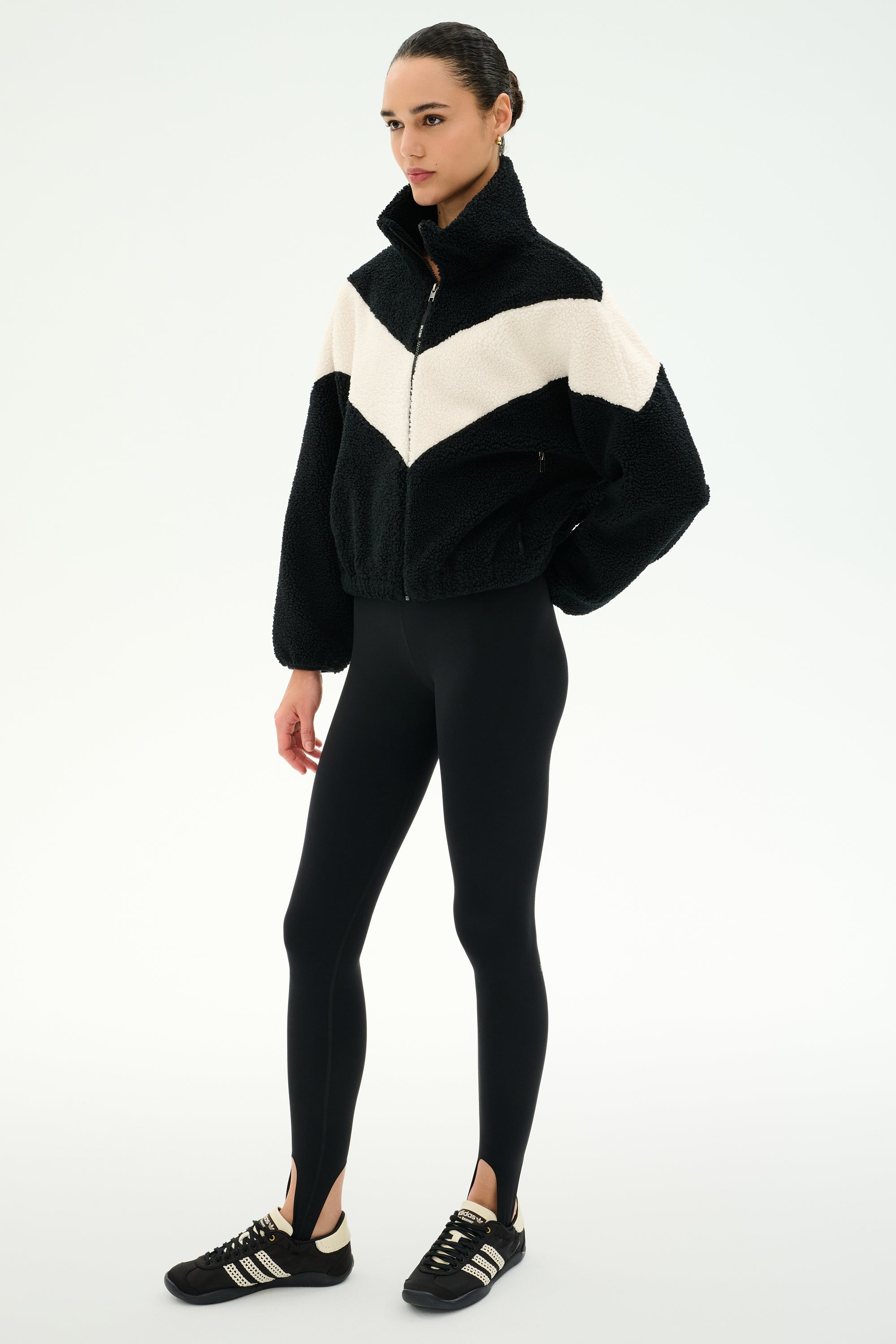 A person stands wearing the SPLITS59 Lottie Chevron Sherpa Full Zip Jacket in Black/Creme, crafted from soft polyester fleece, paired with black leggings with stirrups and black Adidas sneakers against a plain white background.