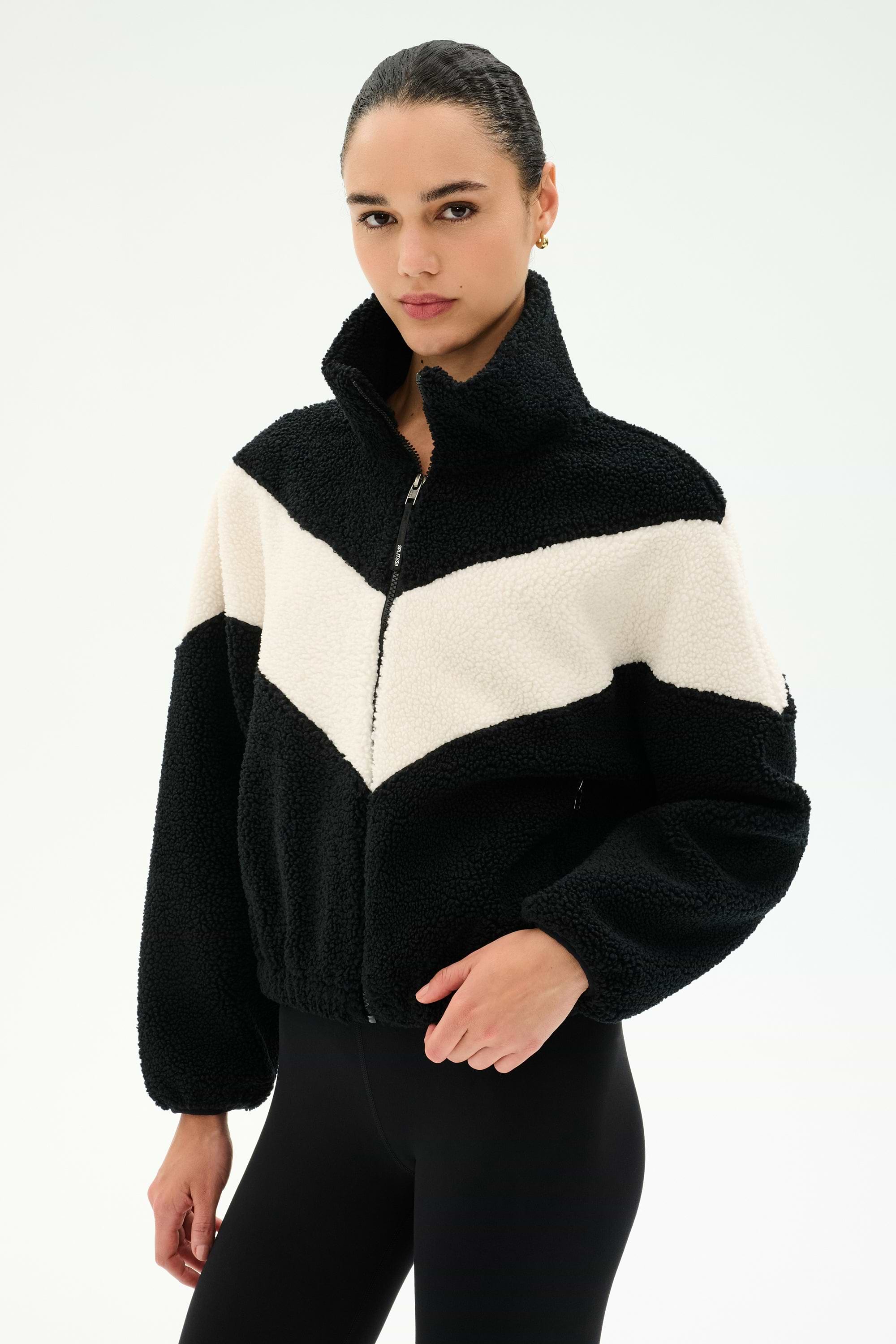 A person wearing the SPLITS59 Lottie Chevron Sherpa Full Zip Jacket in Black/Creme, featuring a chevron pattern and crafted from 100% polyester, stands against a plain light background.