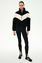 A person stands facing forward against a plain white background, wearing the SPLITS59 Lottie Chevron Sherpa Full Zip Jacket in Black/Creme, crafted from 100% polyester, along with black leggings with stirrups and black Adidas sneakers.