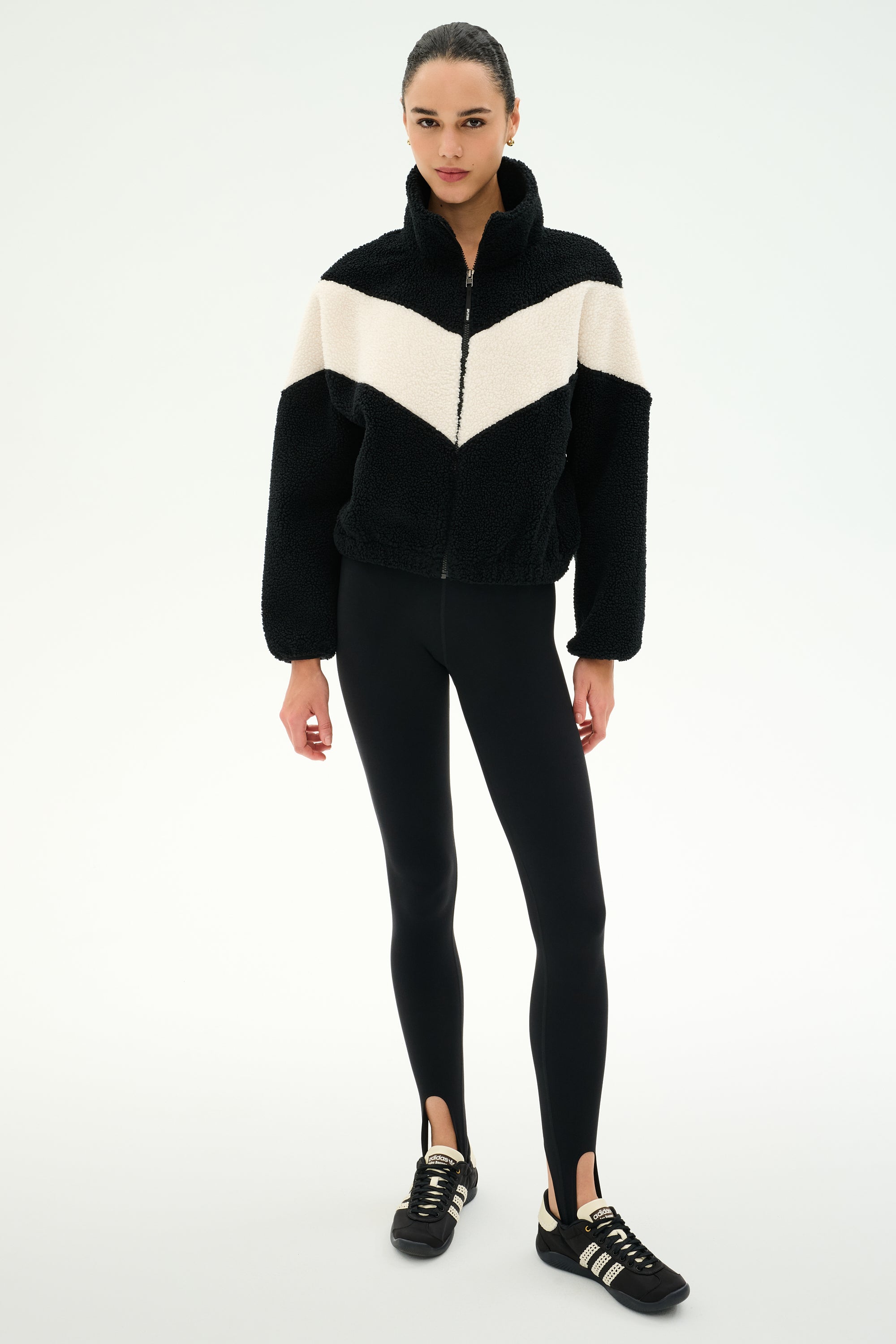 A person stands facing forward against a plain white background, wearing the SPLITS59 Lottie Chevron Sherpa Full Zip Jacket in Black/Creme, crafted from 100% polyester, along with black leggings with stirrups and black Adidas sneakers.