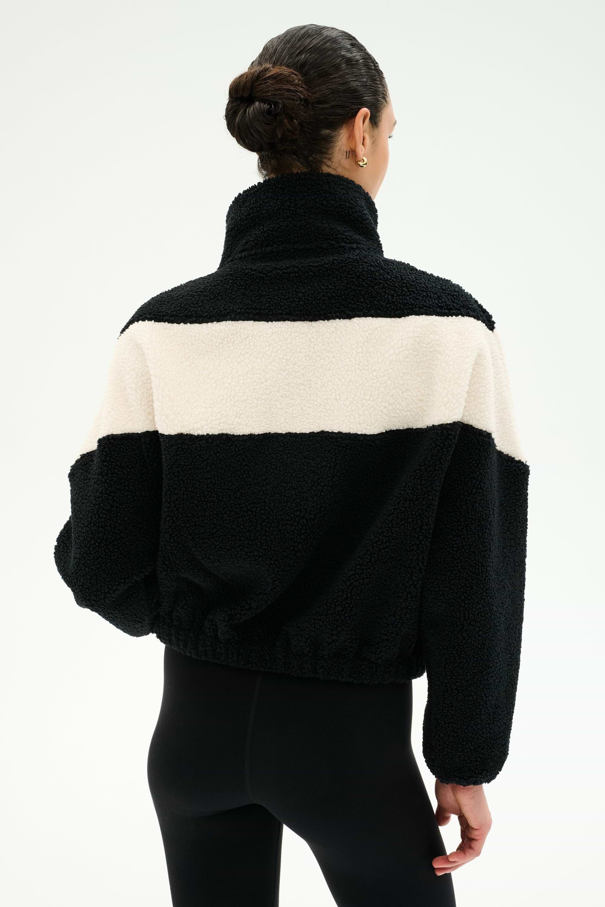 A person with dark hair in a bun is seen from the back against a plain light background, wearing the SPLITS59 Lottie Chevron Sherpa Full Zip Jacket in Black/Creme—crafted from soft 100% polyester—and black leggings.