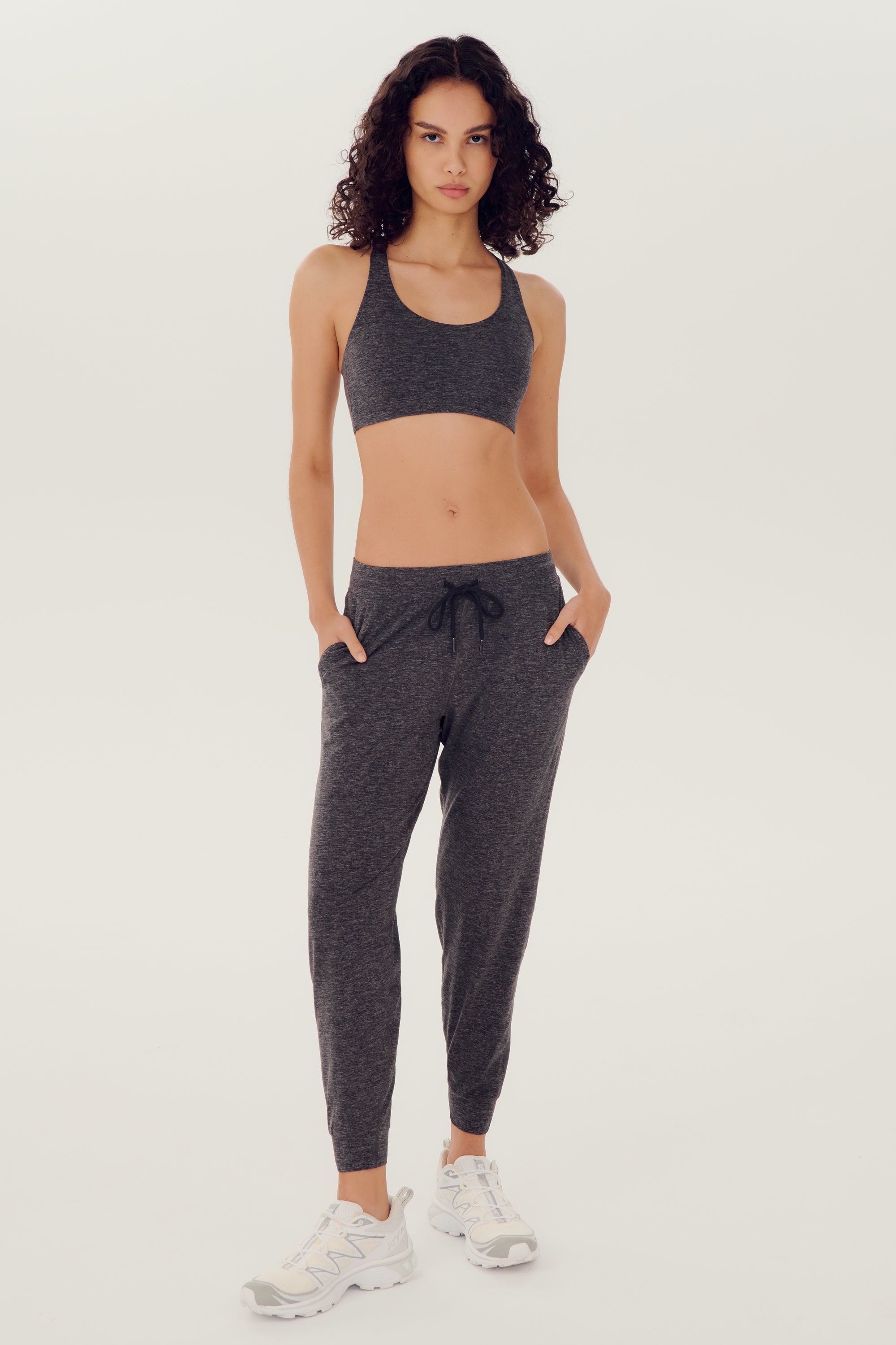 Full front view of woman with dark wavy hair wearing dark grey bra and dark drey joggers with black drawstring paired with white shoes