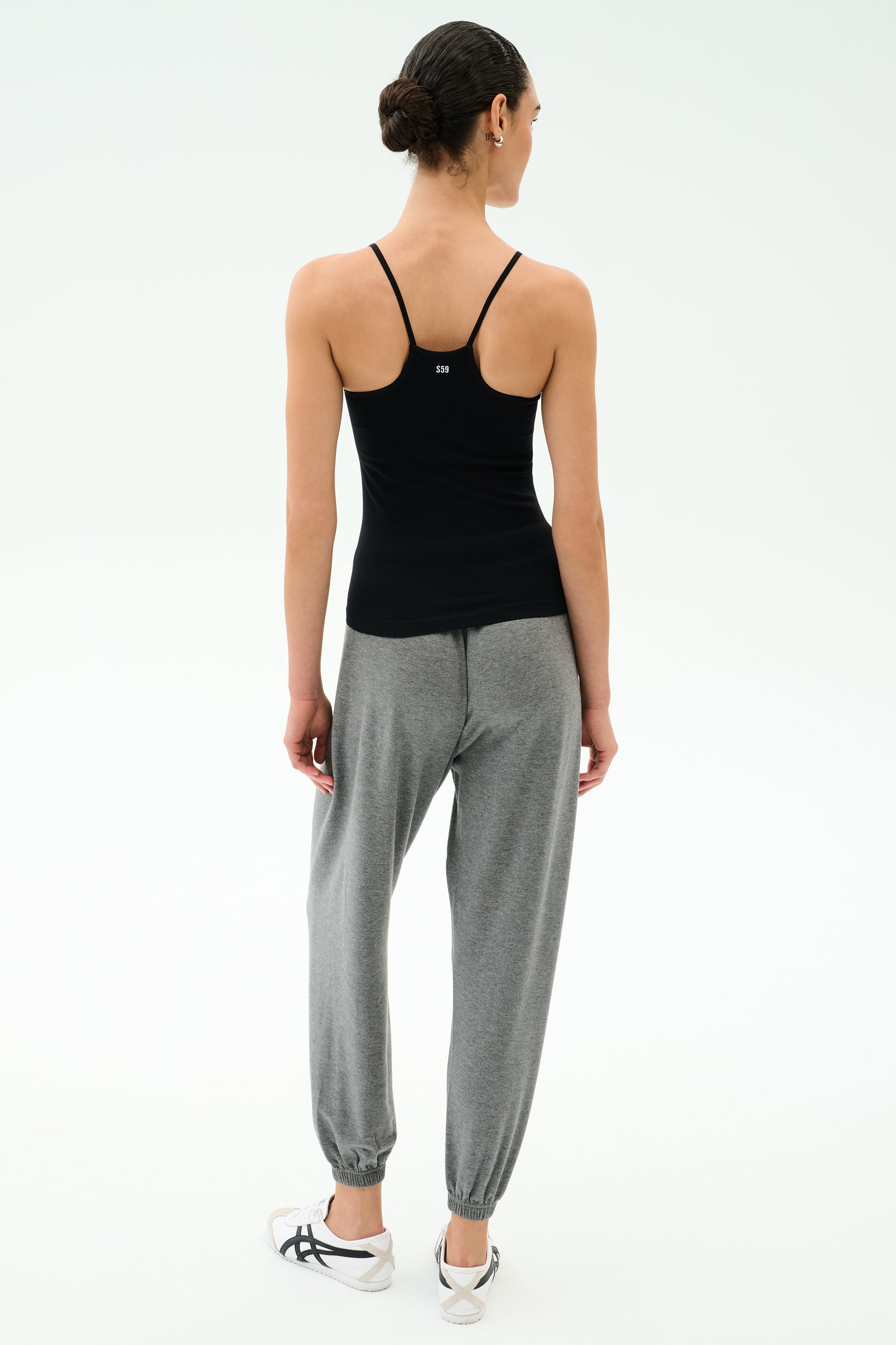 A woman faces away, wearing the SPLITS59 Loren Seamless Waist Length Tank in black, paired with gray jogger pants and white sneakers against a plain light background.