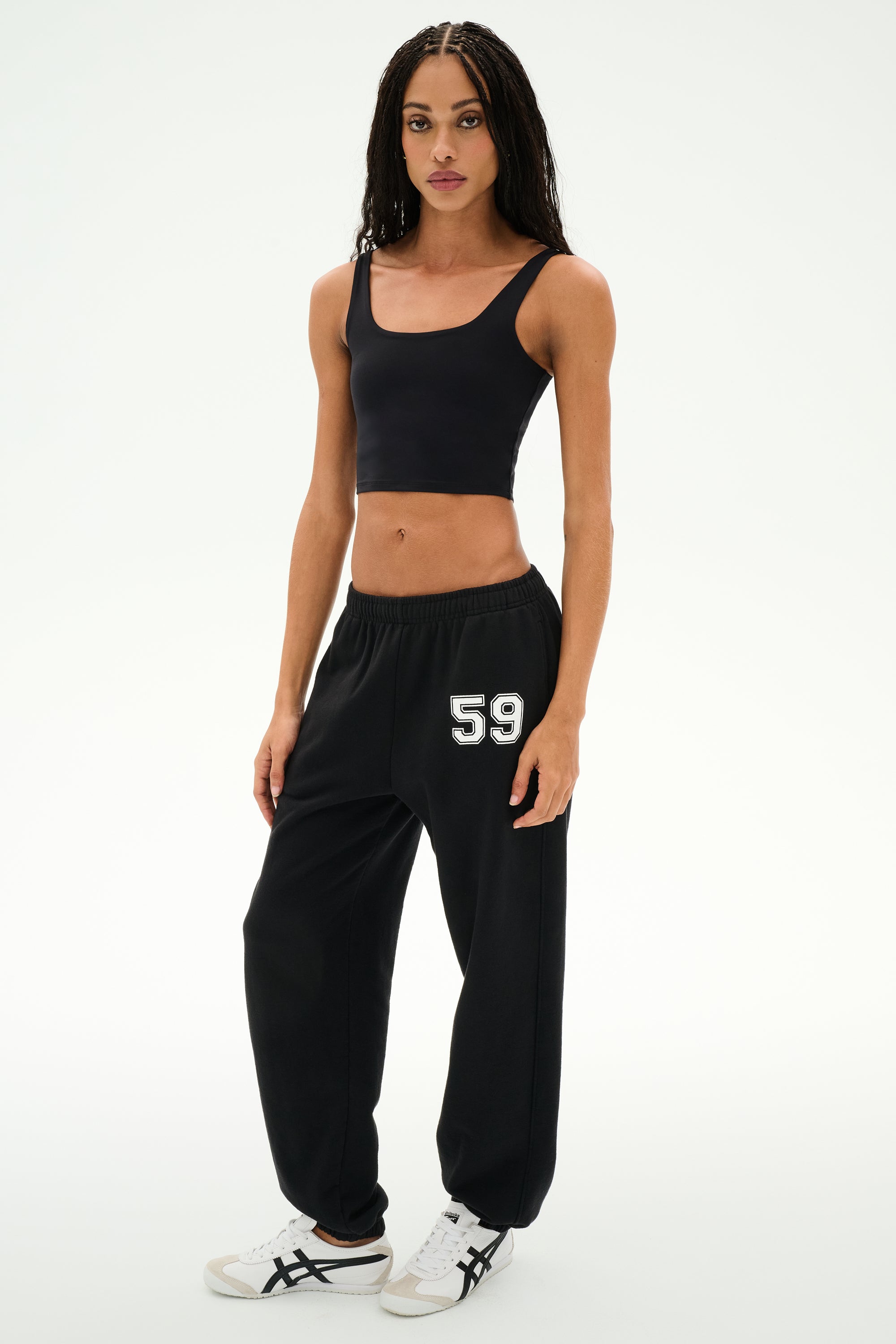 Wearing a black crop top and SPLITS59 Serena French Terry Oversized Sweatpant in Black/59—featuring the “59” detail on the left leg and made in Los Angeles—the person stands against a plain white background, paired with white sneakers.