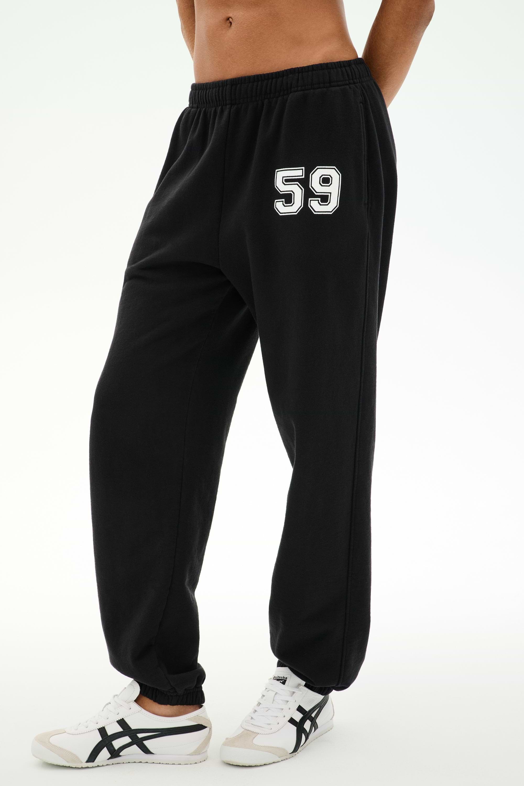 Model wears the SPLITS59 Serena French Terry Oversized Sweatpant in Black/59, featuring the number 59 printed on the left thigh. Made in Los Angeles for a comfortable and stylish look, these sweatpants are paired with white sneakers with black stripes and set against a plain background.