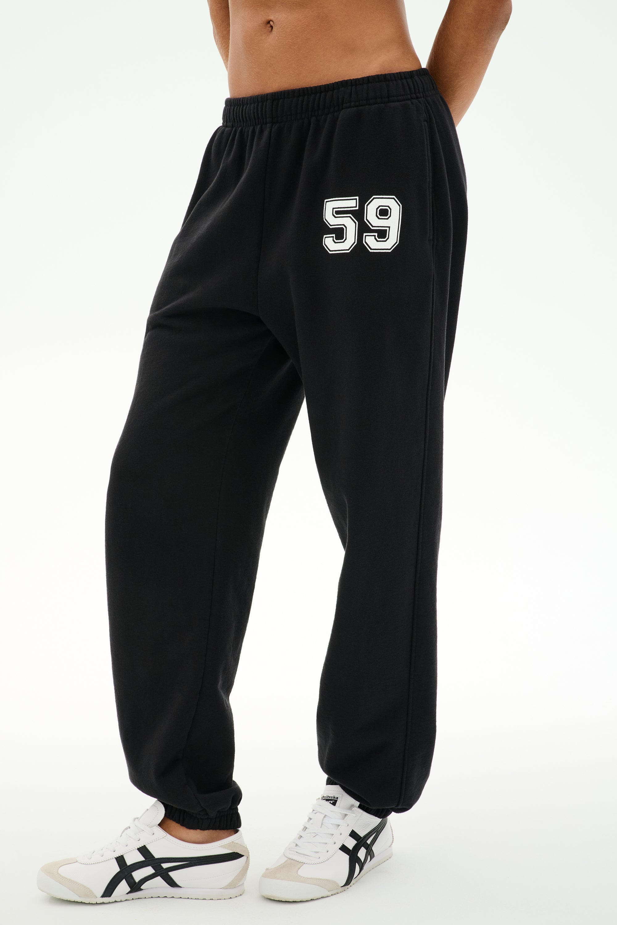 Model wears the SPLITS59 Serena French Terry Oversized Sweatpant in Black/59, featuring the number 59 printed on the left thigh. Made in Los Angeles for a comfortable and stylish look, these sweatpants are paired with white sneakers with black stripes and set against a plain background.
