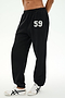 Model wears the SPLITS59 Serena French Terry Oversized Sweatpant in Black/59, featuring the number 59 printed on the left thigh. Made in Los Angeles for a comfortable and stylish look, these sweatpants are paired with white sneakers with black stripes and set against a plain background.