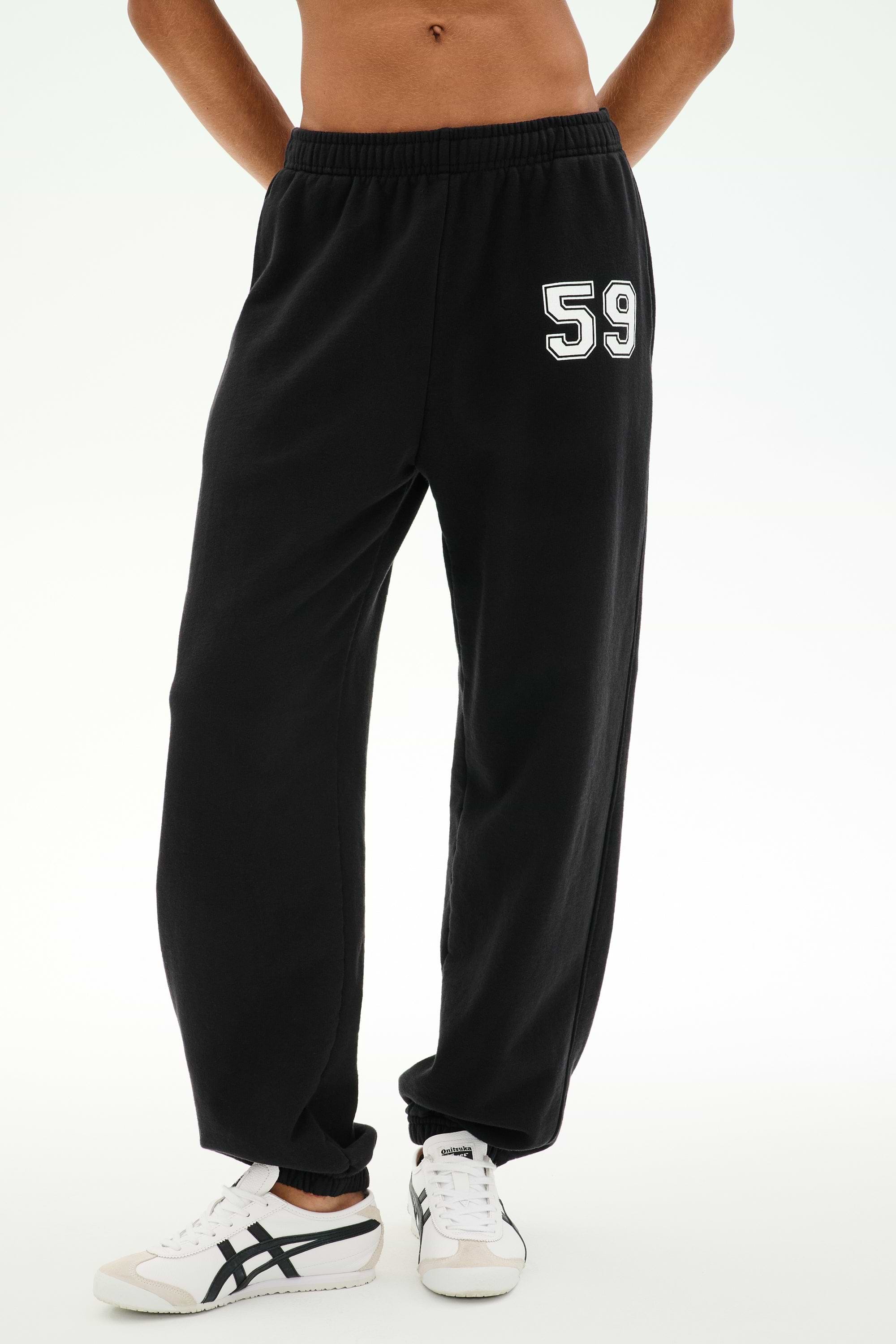 A person is wearing the SPLITS59 Serena French Terry Oversized Sweatpant in black with a “59” on the left thigh, paired with white sneakers featuring black accents, standing against a plain background; these sweatpants are made in Los Angeles.