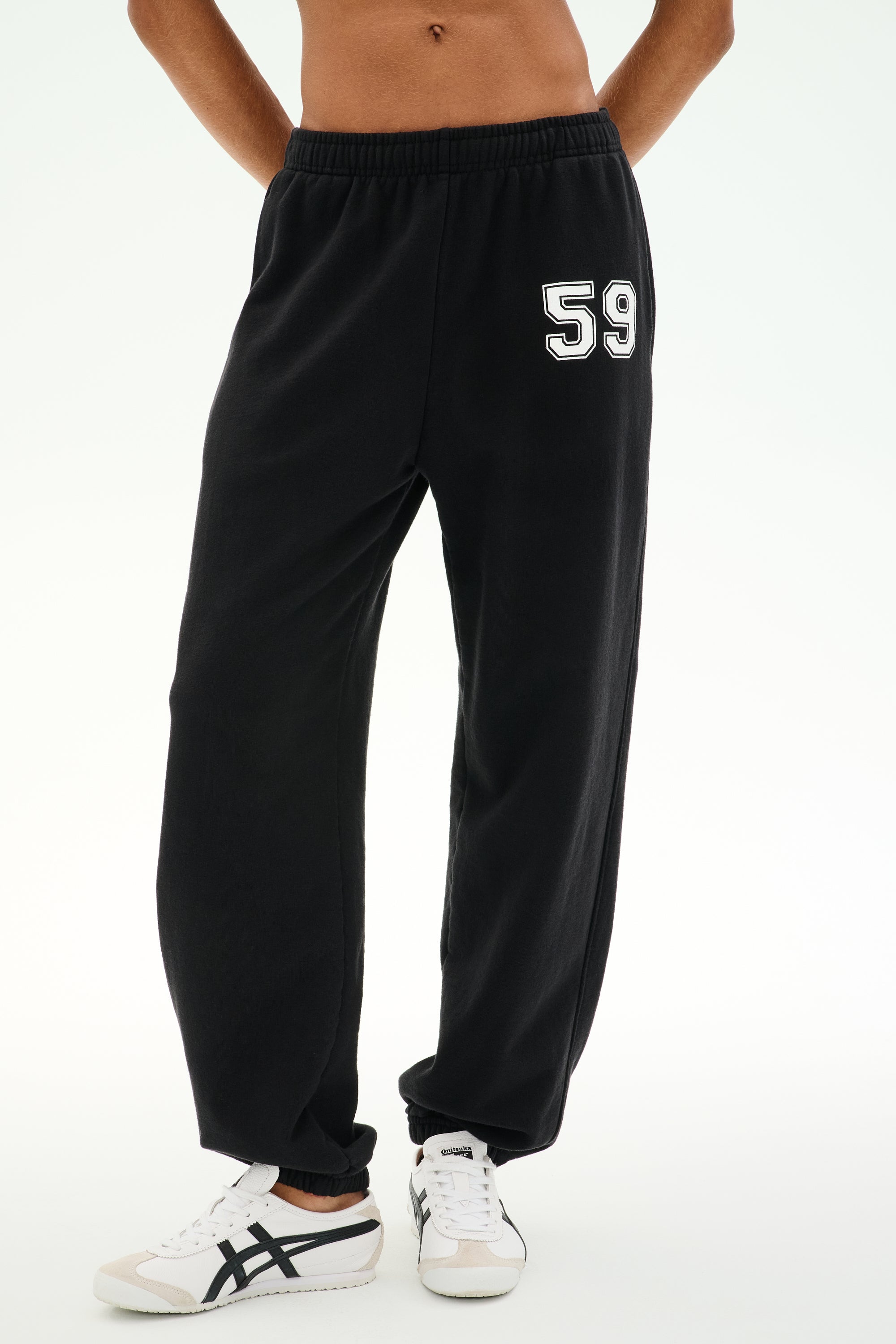 A person is wearing the SPLITS59 Serena French Terry Oversized Sweatpant in black with a “59” on the left thigh, paired with white sneakers featuring black accents, standing against a plain background; these sweatpants are made in Los Angeles.
