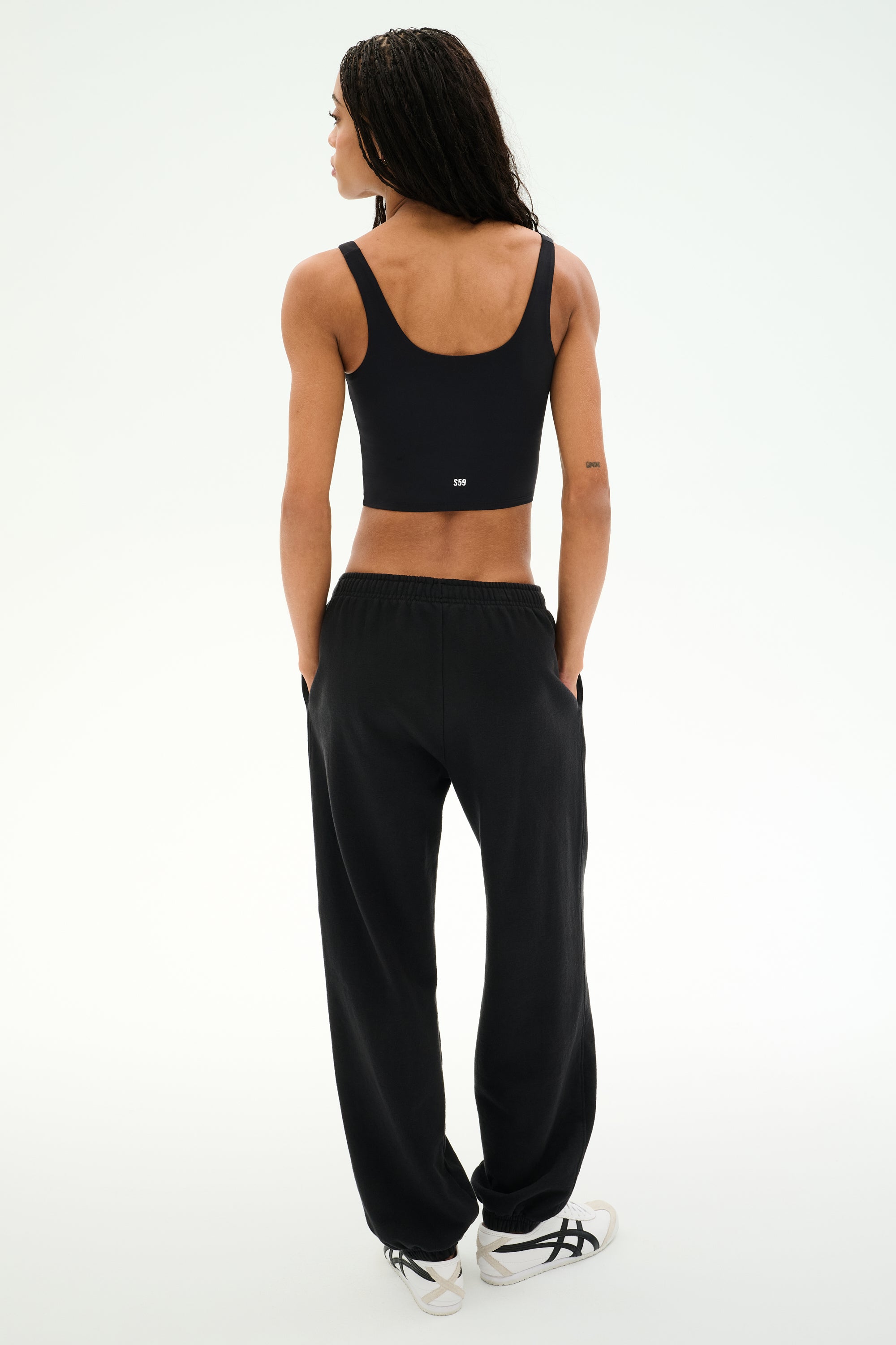 A person stands facing away with hands in pockets against a plain light background, wearing a black crop top, white sneakers, and the SPLITS59 Serena French Terry Oversized Sweatpant in Black/59, made in Los Angeles.