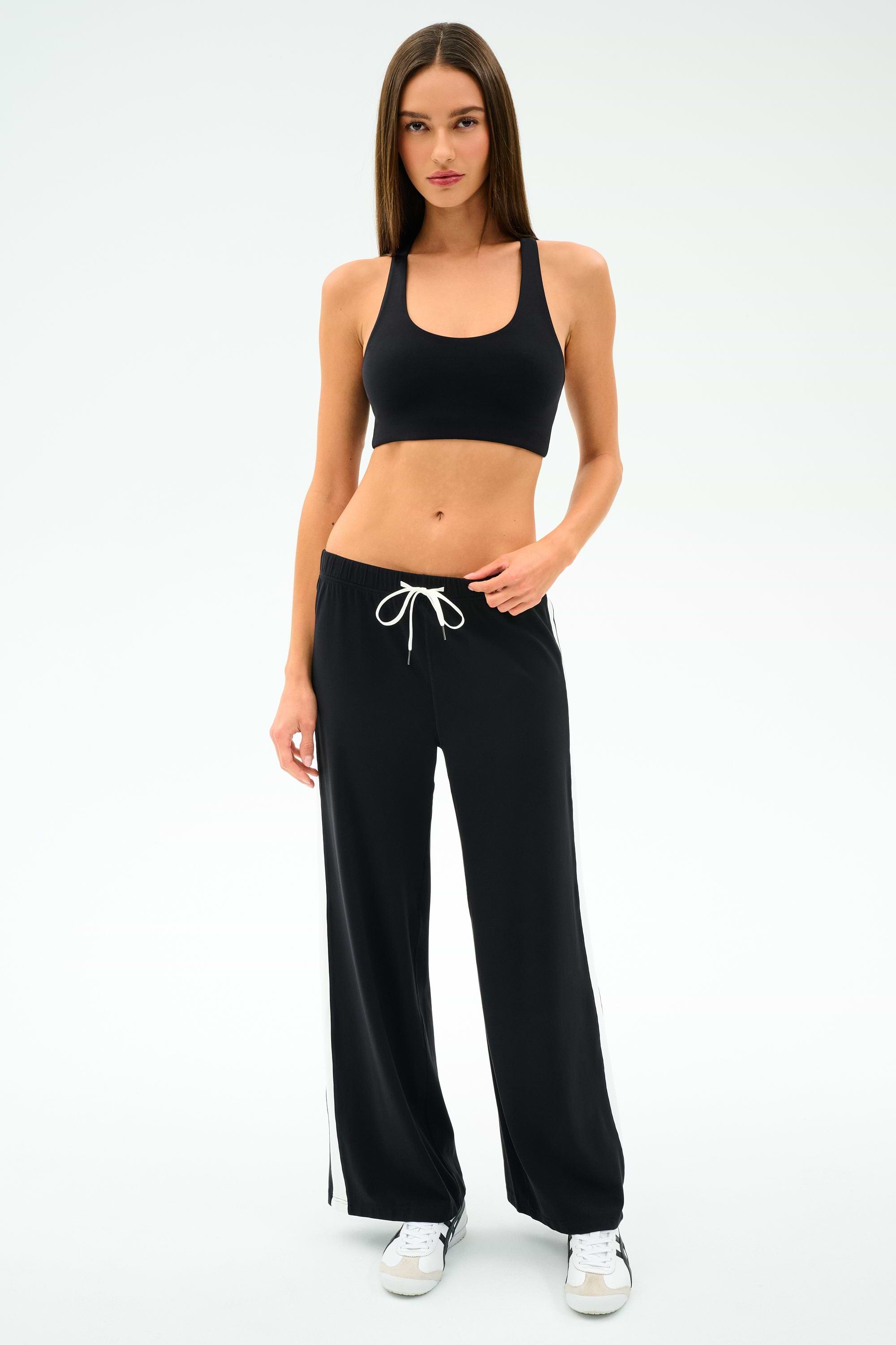 A woman wearing the SPLITS59 Willow Airweight Wide Leg pants in Black/White with a black sports bra and white sneakers stands against a white background.