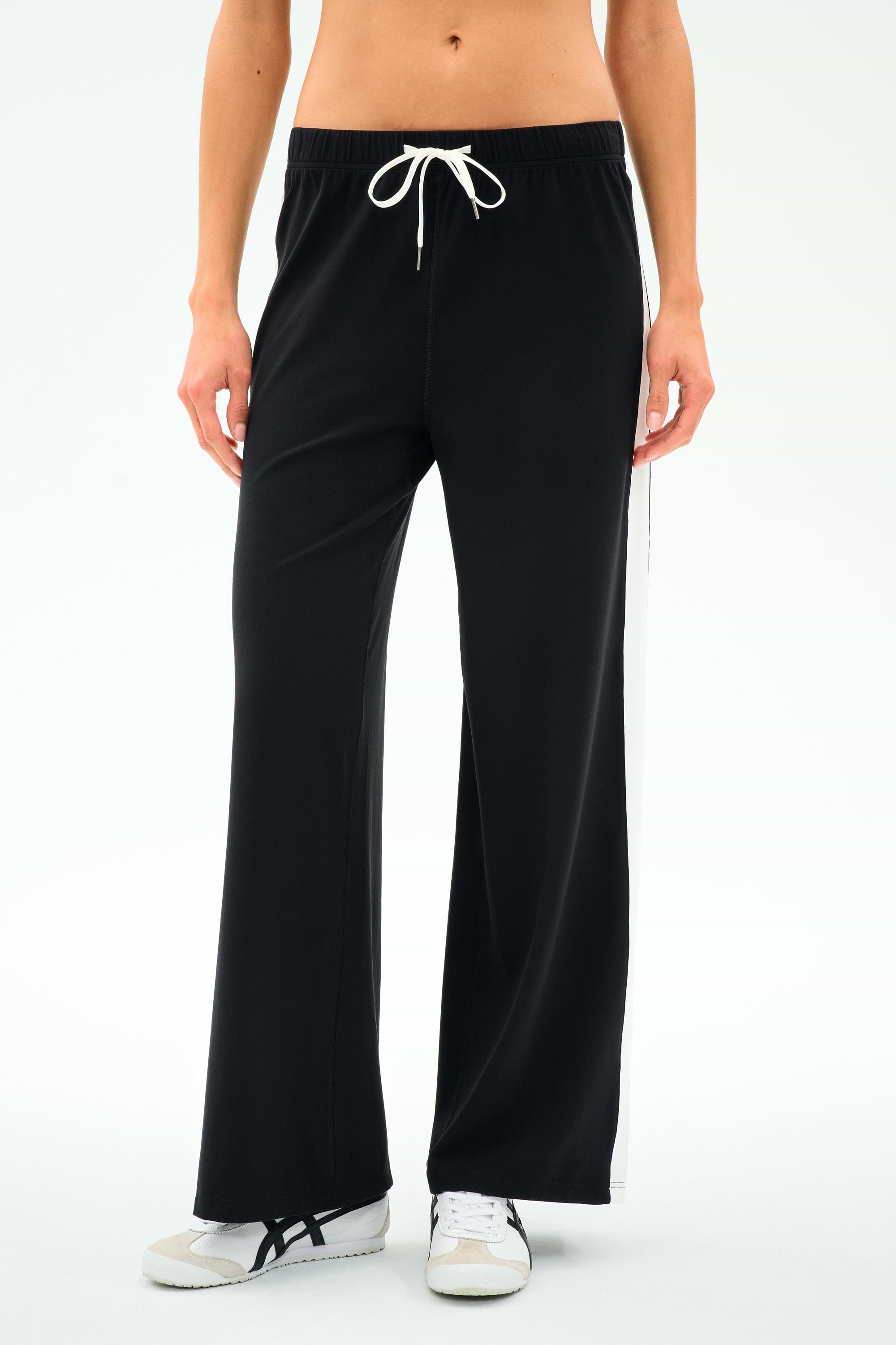A person wearing SPLITS59's Willow Airweight Wide Leg pants in black with white side stripes and white sneakers, standing against a plain white background.