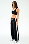 A woman stands against a white background wearing SPLITS59’s Willow Airweight Wide Leg - Black/White pants and a black sports bra with white sneakers—perfect for Pilates.