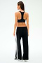 A woman faces away, wearing SPLITS59's Willow Airweight Wide Leg pants in Black/White with white side stripes, a black sports bra, and white sneakers—ideal for Pilates or hot yoga—set against a simple light backdrop.