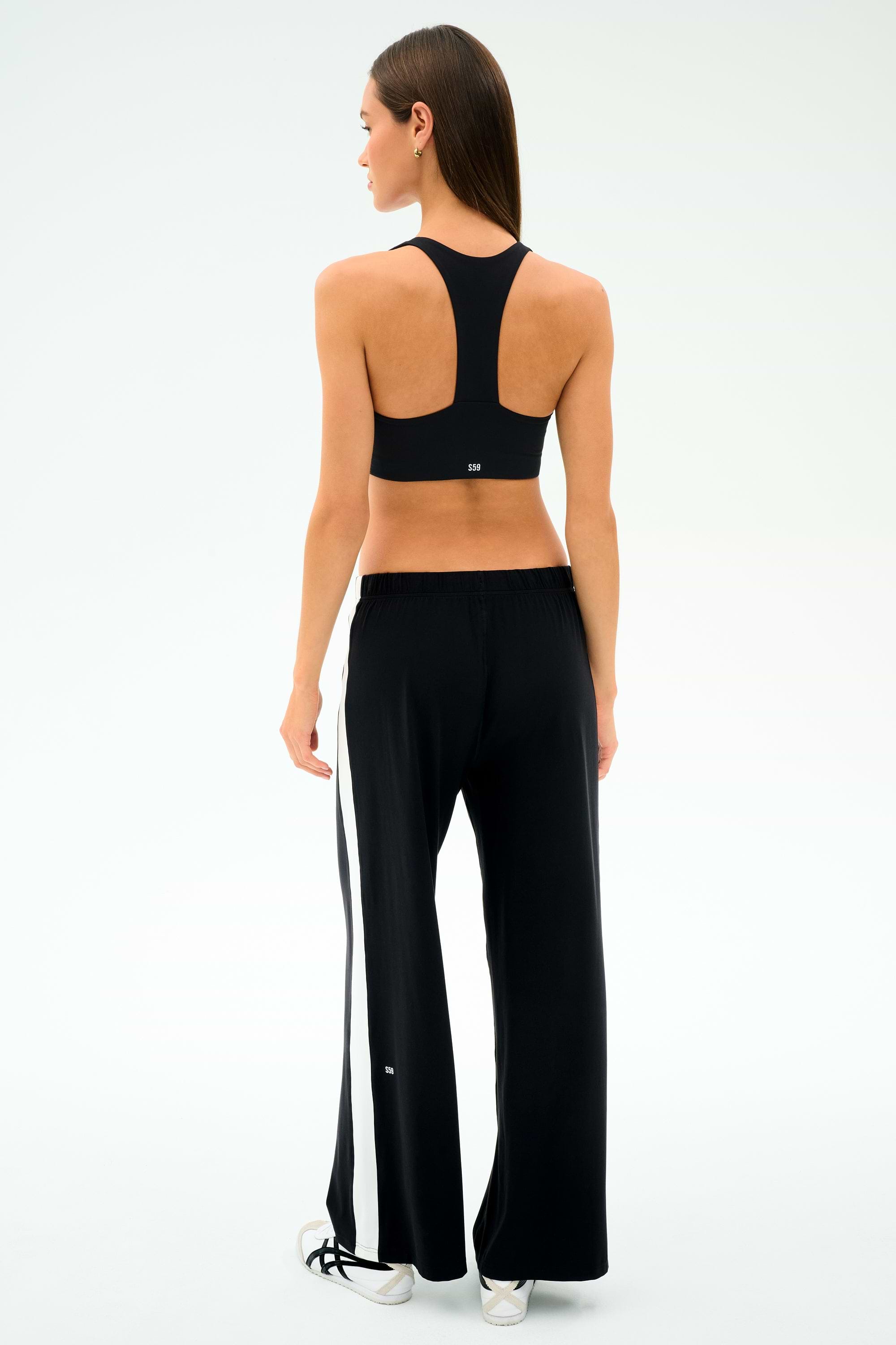 A woman faces away, wearing SPLITS59's Willow Airweight Wide Leg pants in Black/White with white side stripes, a black sports bra, and white sneakers—ideal for Pilates or hot yoga—set against a simple light backdrop.