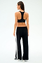 A woman faces away, wearing SPLITS59's Willow Airweight Wide Leg pants in Black/White with white side stripes, a black sports bra, and white sneakers—ideal for Pilates or hot yoga—set against a simple light backdrop.