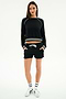 A woman faces forward wearing a black long-sleeve top with striped cuffs and hem, stretchy fabric, SPLITS59 Reena Fleece Sweatshort in black, white socks, and white sneakers.