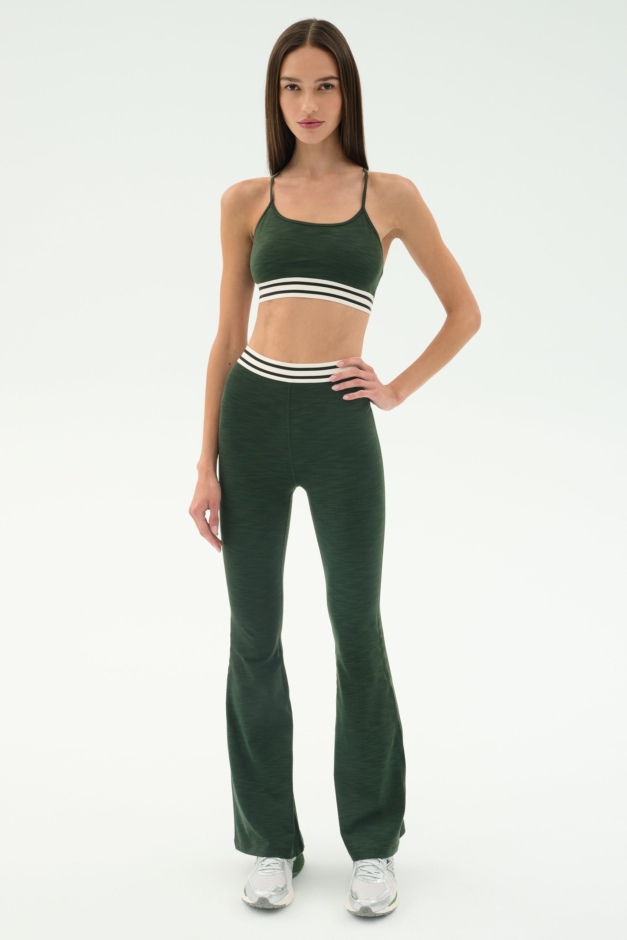 A woman stands in a studio wearing the SPLITS59 Cali Active Rib Bra in Heather Military with matching high-waisted leggings featuring white stripes, paired with white sneakers.