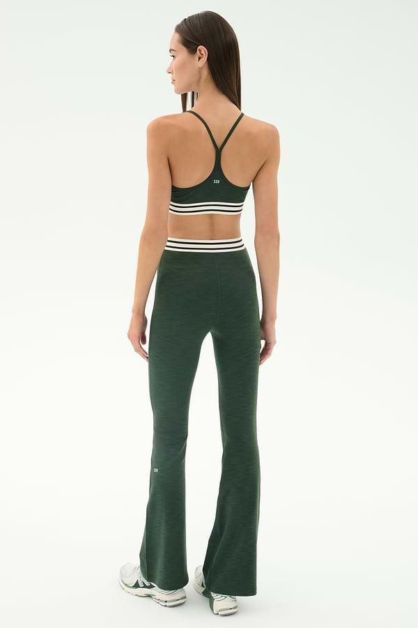 A woman stands facing away in the SPLITS59 Cali Active Rib Bra - Heather Military, paired with matching flared leggings featuring white stripes and white sneakers, set against a plain light background.