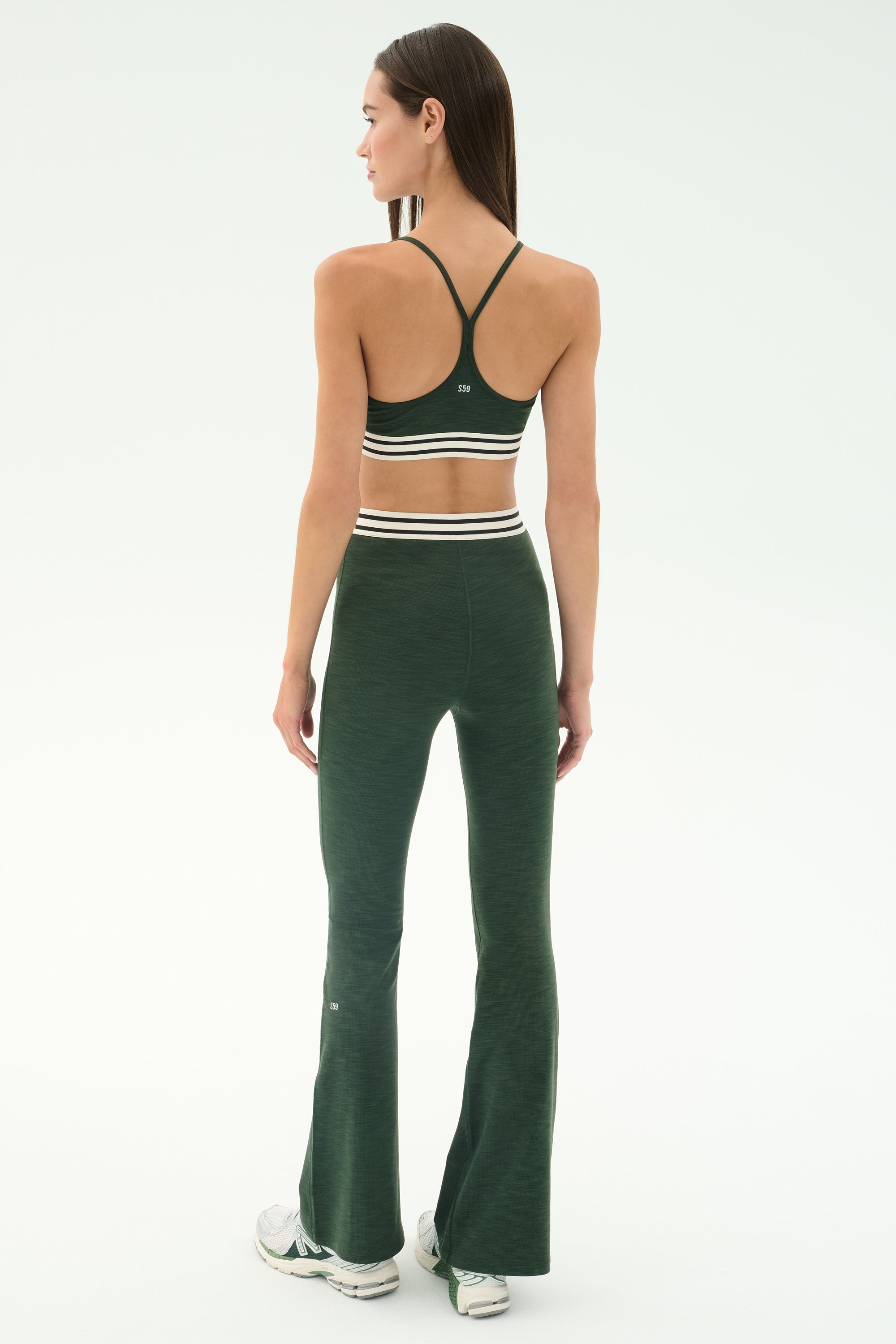 A woman stands facing away in the SPLITS59 Cali Active Rib Bra - Heather Military, paired with matching flared leggings featuring white stripes and white sneakers, set against a plain light background.