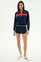 A woman wears the SPLITS59 Carrie Woven Nylon Jacket in Indigo/Lotus Red, paired with matching shorts and white sneakers with red stripes, standing against a plain white background.