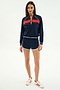 A woman wears the SPLITS59 Carrie Woven Nylon Jacket in Indigo/Lotus Red, paired with matching shorts and white sneakers with red stripes, standing against a plain white background.
