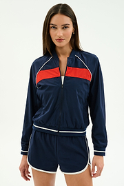 A woman faces forward wearing the SPLITS59 Carrie Woven Nylon Jacket in Indigo/Lotus Red, paired with matching navy blue spandex shorts featuring white trim.