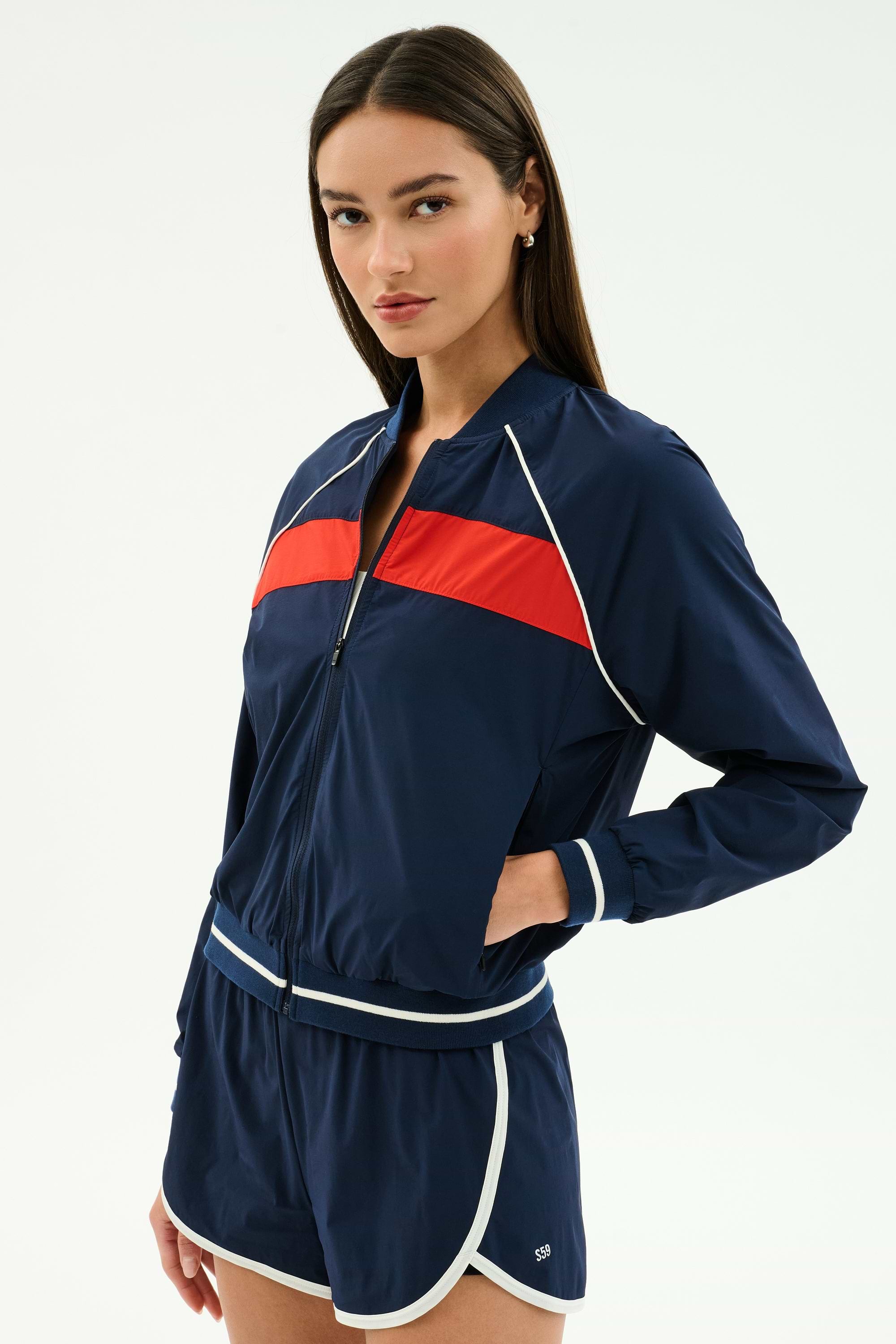A woman wears the SPLITS59 Carrie Woven Nylon Jacket in Indigo/Lotus Red with matching nylon shorts featuring white trim, standing with one hand in her pocket.