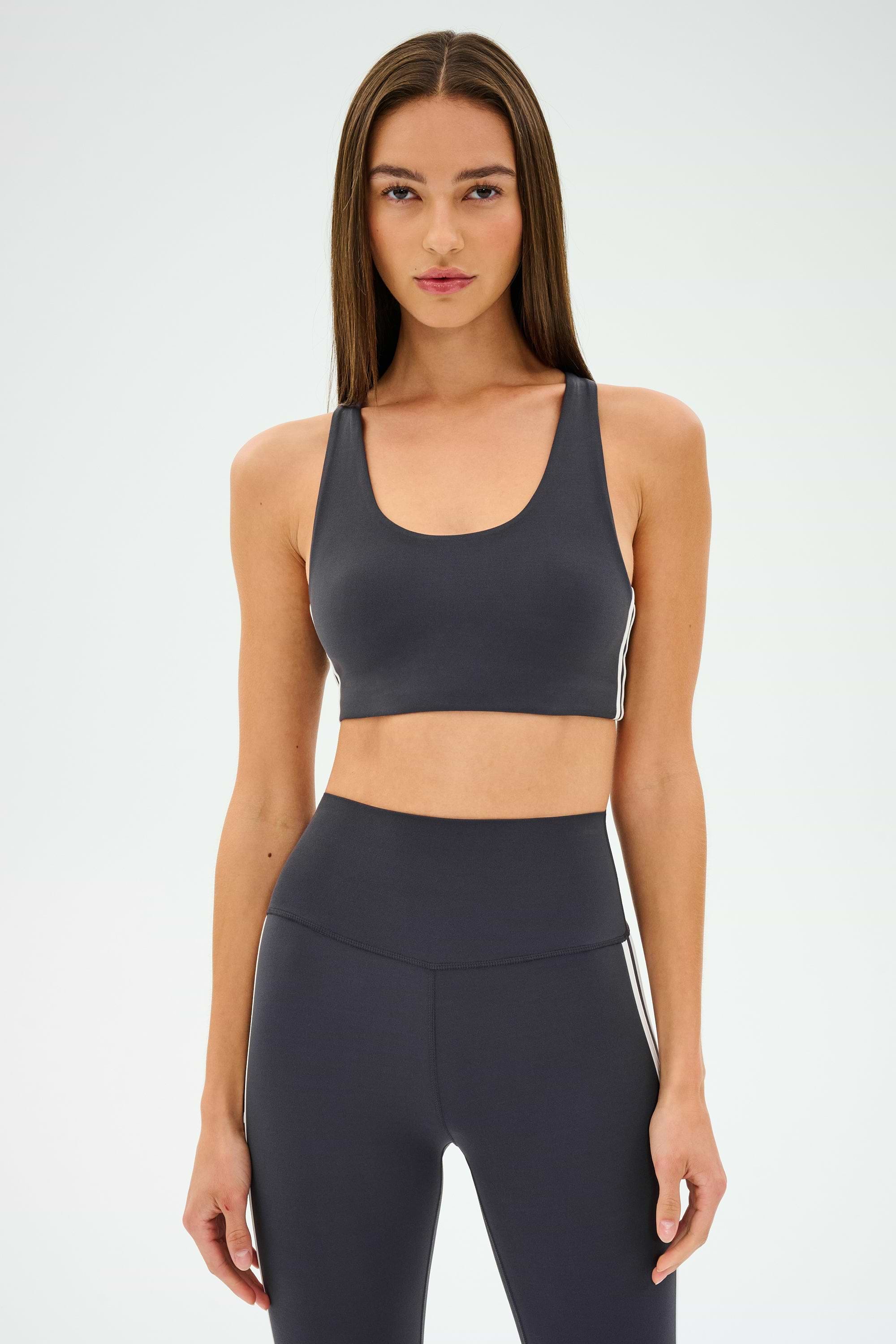 A person wearing an Ella Airweight Bra in Graphite/White from SPLITS59 stands against a plain background, ready for hot yoga.