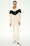 A woman stands against a plain white background wearing the SPLITS59 Lottie Chevron Sherpa Full Zip Jacket in Creme/Black, featuring a black chevron design and crafted from smooth polyester, paired with cream pants with black stripes and cream sneakers accented with black stripes.