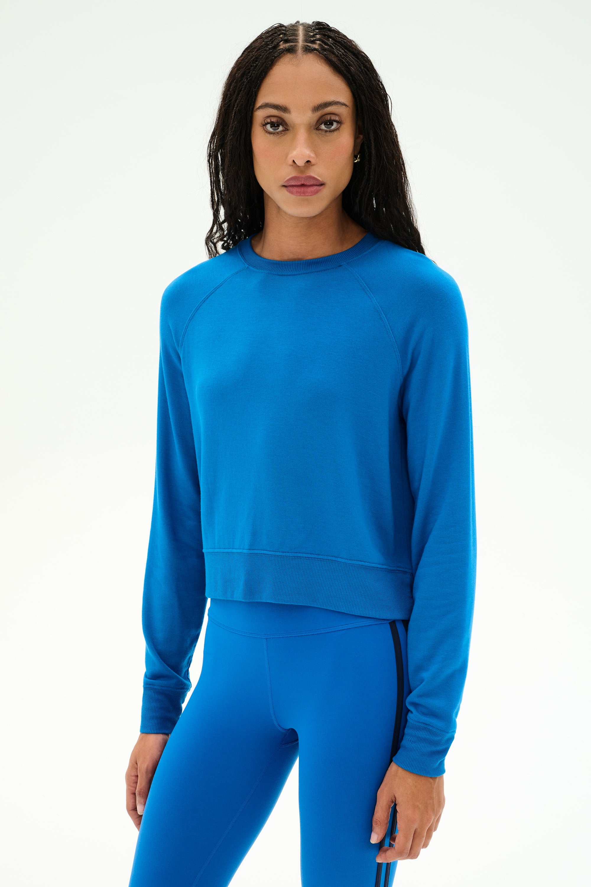 A woman with long dark hair stands against a plain white background wearing the SPLITS59 Brooks Fleece Long Sleeve Sweatshirt in Classic Blue, paired with matching blue leggings, both crafted from a soft modal spandex blend.