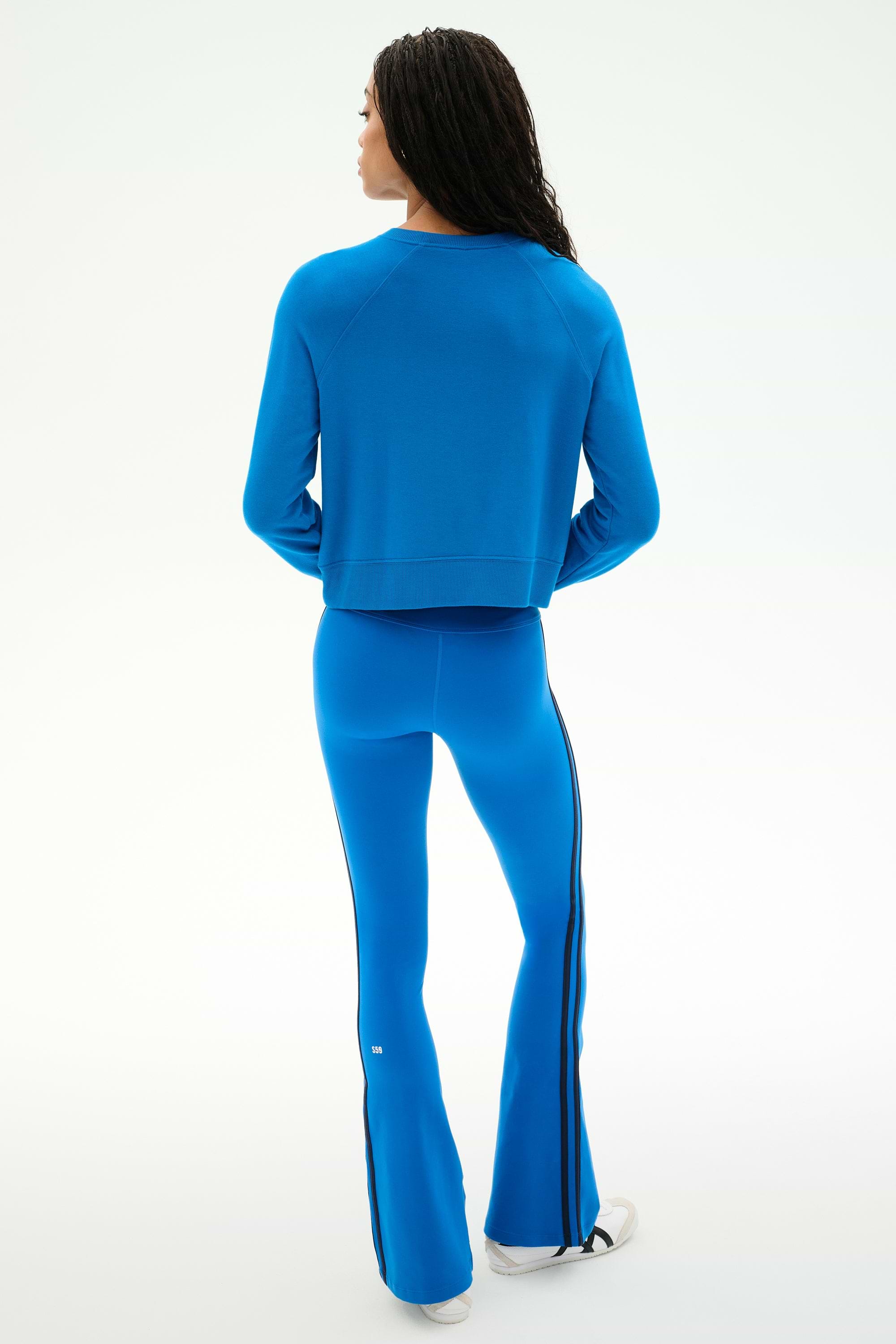 A person stands with their back to the camera, dressed in the SPLITS59 Brooks Fleece Long Sleeve Sweatshirt in Classic Blue and matching blue flared pants with black side stripes, crafted from a soft modal spandex blend, paired with white shoes against a plain light background.