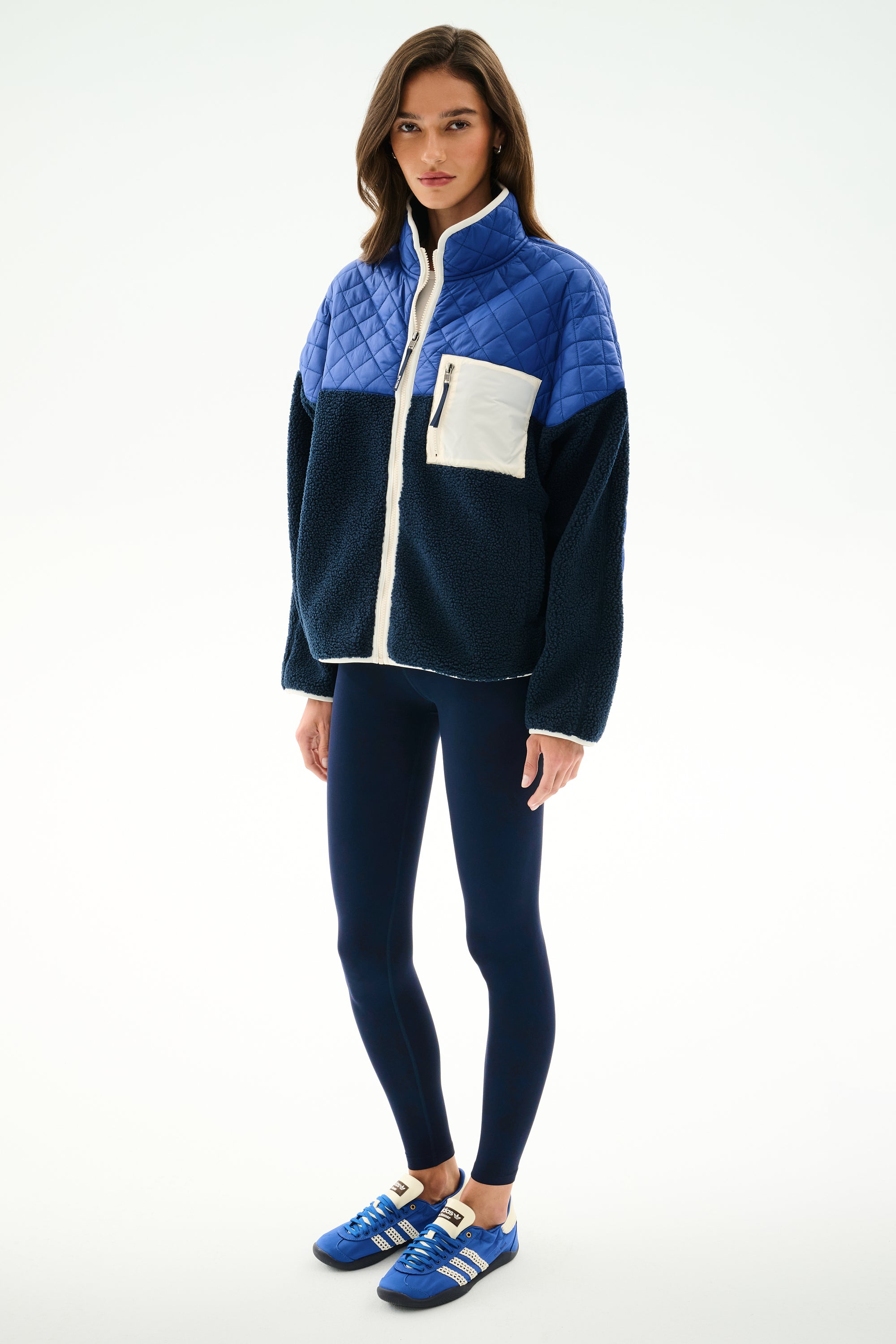 A woman stands in the SPLITS59 Skylar Sherpa Jacket - Indigo/Cobalt, featuring a white pocket, navy leggings, and blue sneakers against a white background.