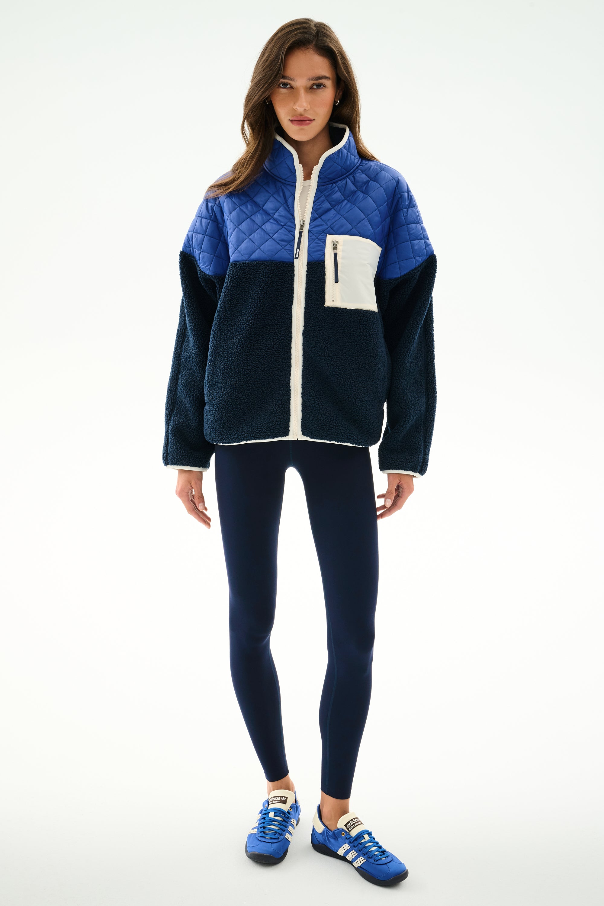 A woman faces forward wearing the SPLITS59 Skylar Sherpa Jacket - Indigo/Cobalt, navy leggings, and blue Adidas sneakers against a plain white background.