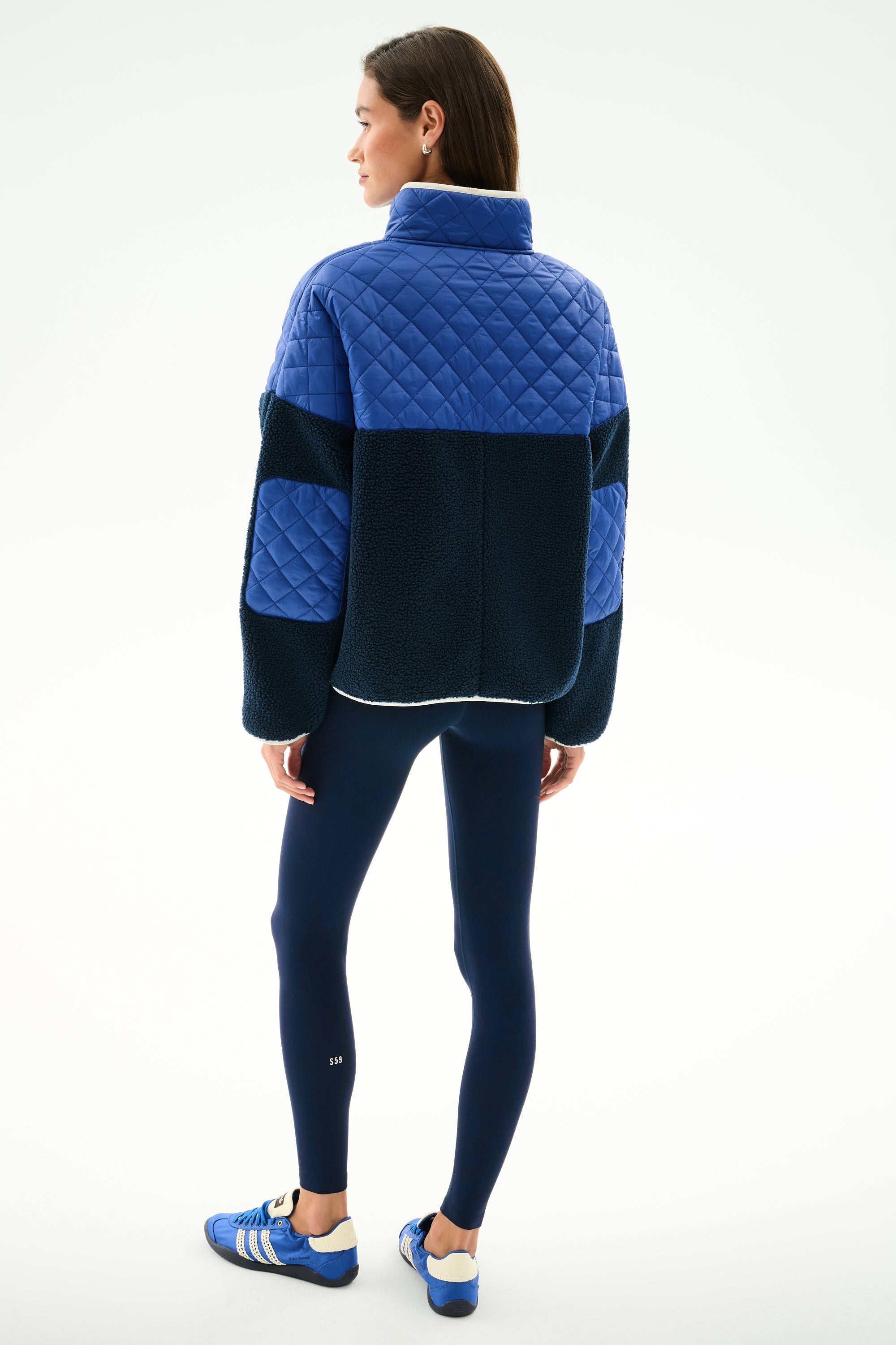 Woman faces away, wearing the SPLITS59 Skylar Sherpa Jacket - Indigo/Cobalt with navy leggings and blue Adidas sneakers, standing against a white background.