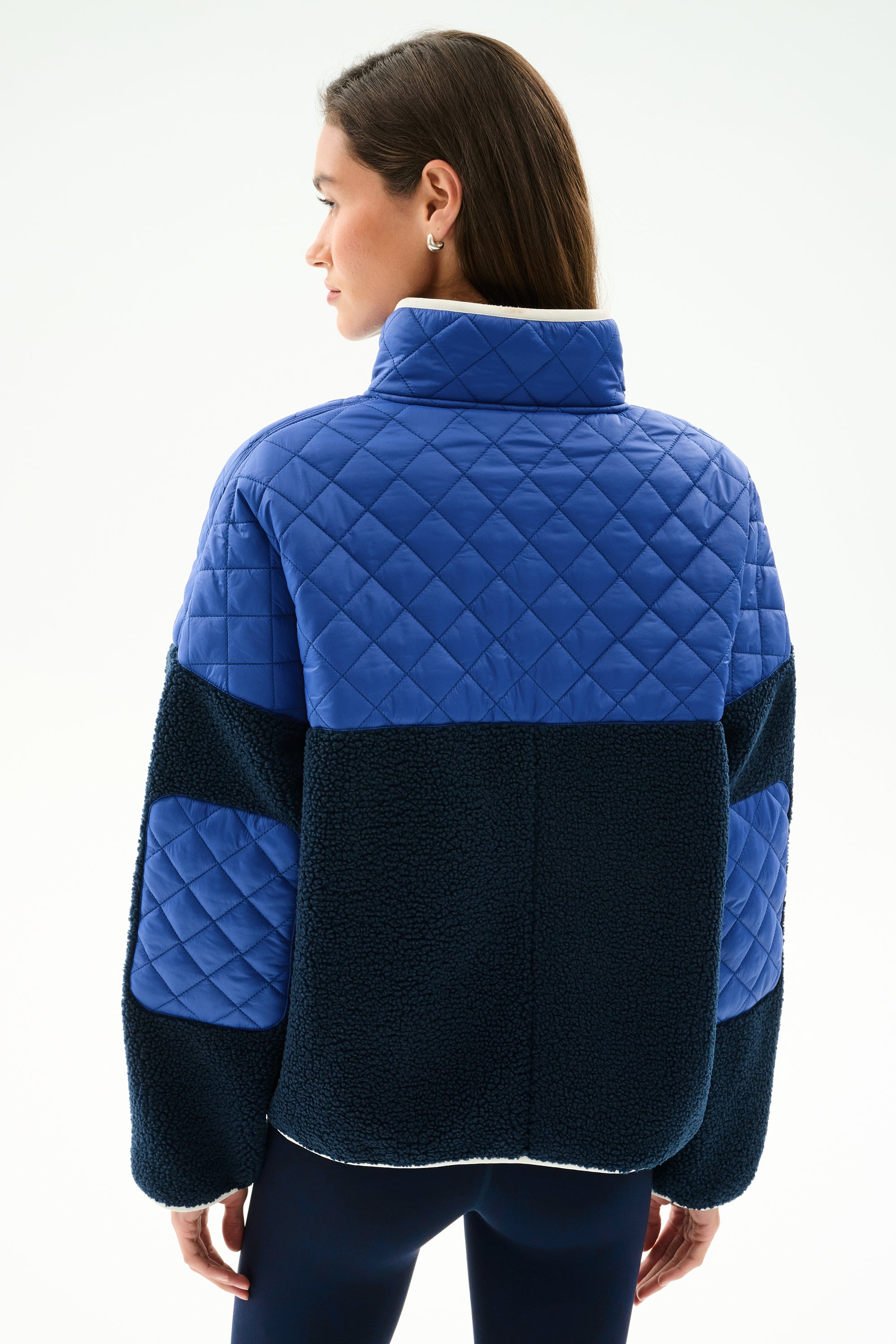A woman with straight brown hair models the SPLITS59 Skylar Sherpa Jacket - Indigo/Cobalt from the back, featuring blue quilted fabric and black panels, against a white background.