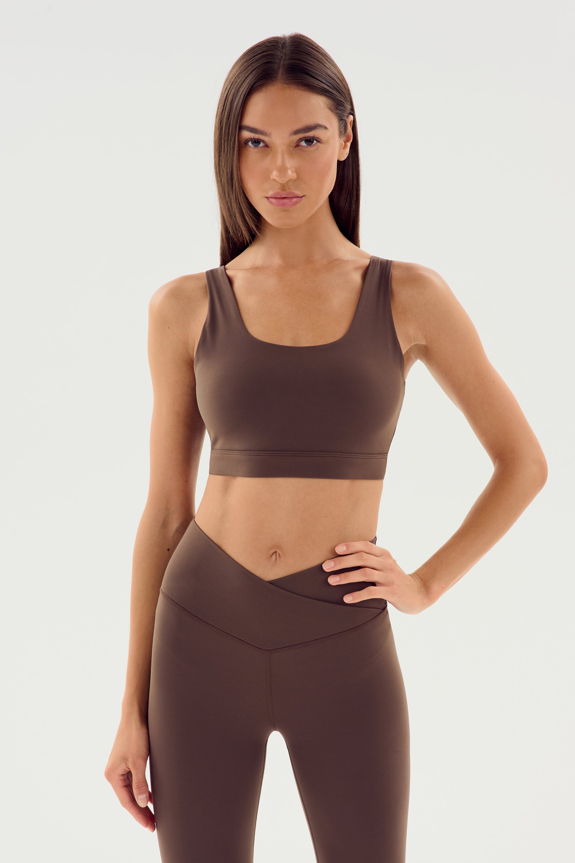 A woman with long hair is standing against a plain background, one hand on her hip, wearing a dark chocolate Sprint Rigor Bra and matching leggings by SPLITS59. Perfect for multi-sport performance and gym workouts.