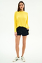 A woman faces forward in the SPLITS59 Andie Oversized Fleece Sweatshirt in bright yellow, paired with black shorts and white sneakers with green stripes, against a plain white background.