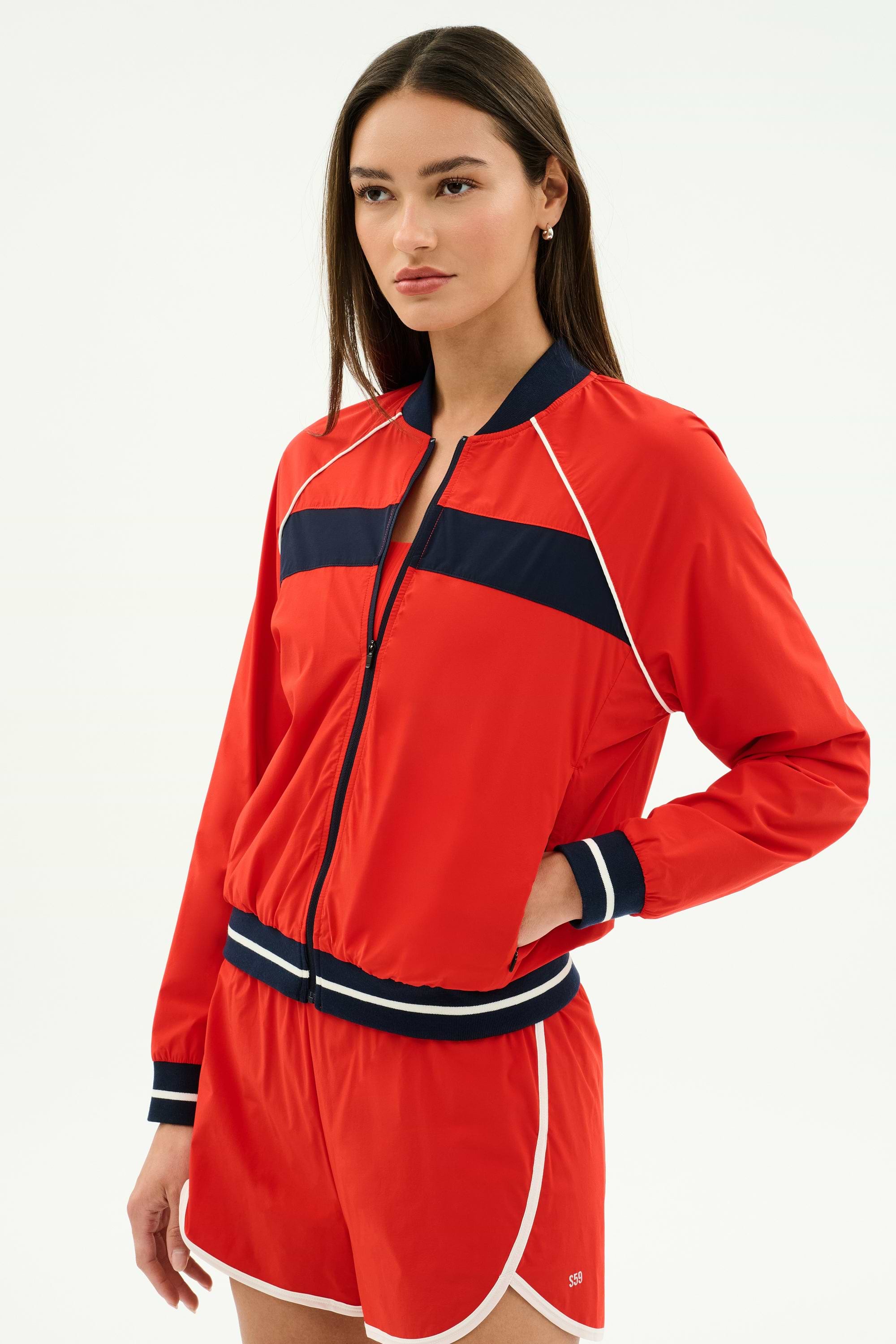 A woman wears the SPLITS59 Carrie Woven Nylon Jacket in Lotus Red/Indigo, paired with matching red shorts, standing against a plain white background.