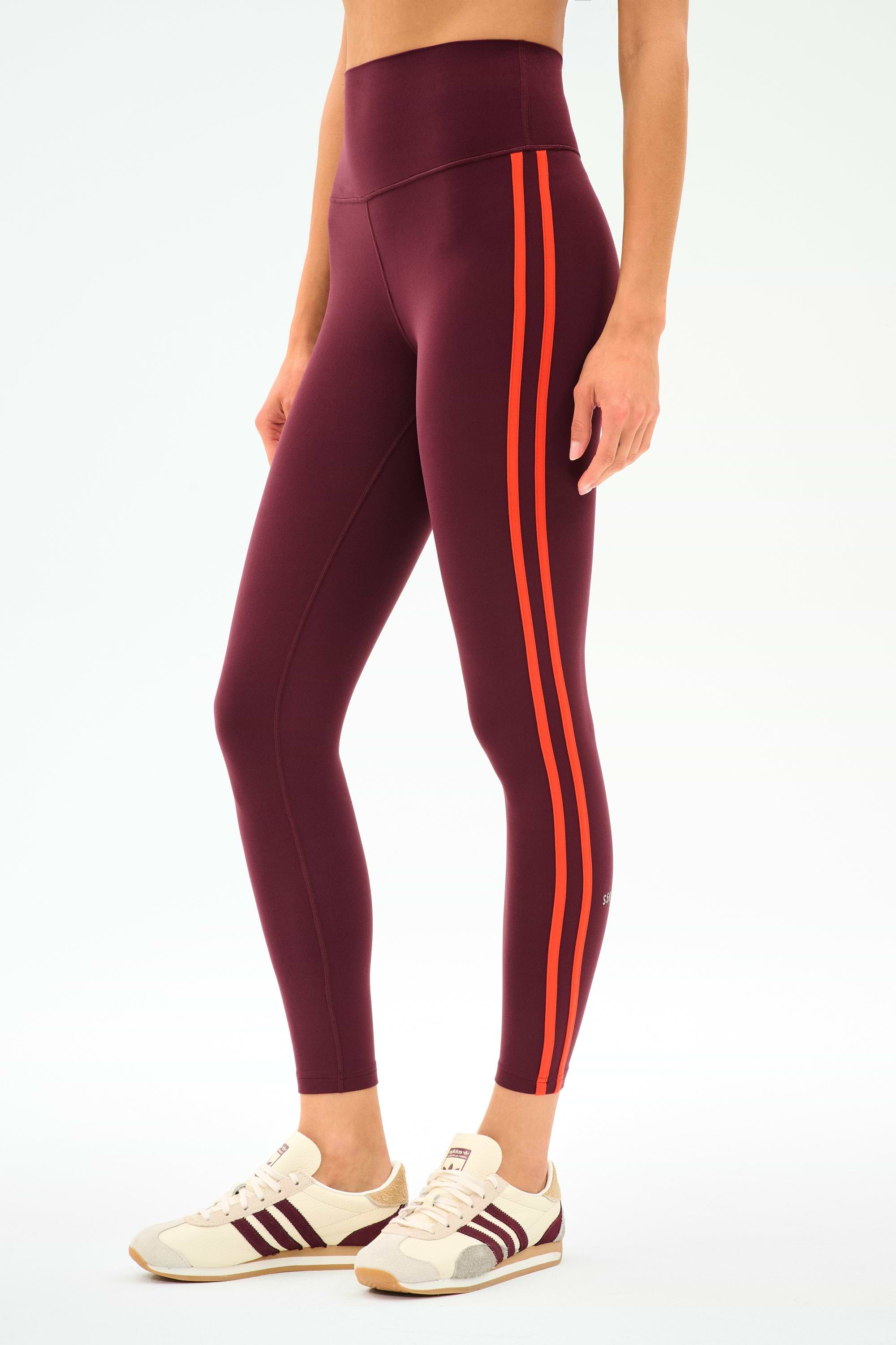 A person wears SPLITS59's Ella High Waist Airweight 7/8 leggings in Dark Cherry/Valencia with white sneakers featuring burgundy accents, standing against a plain background.