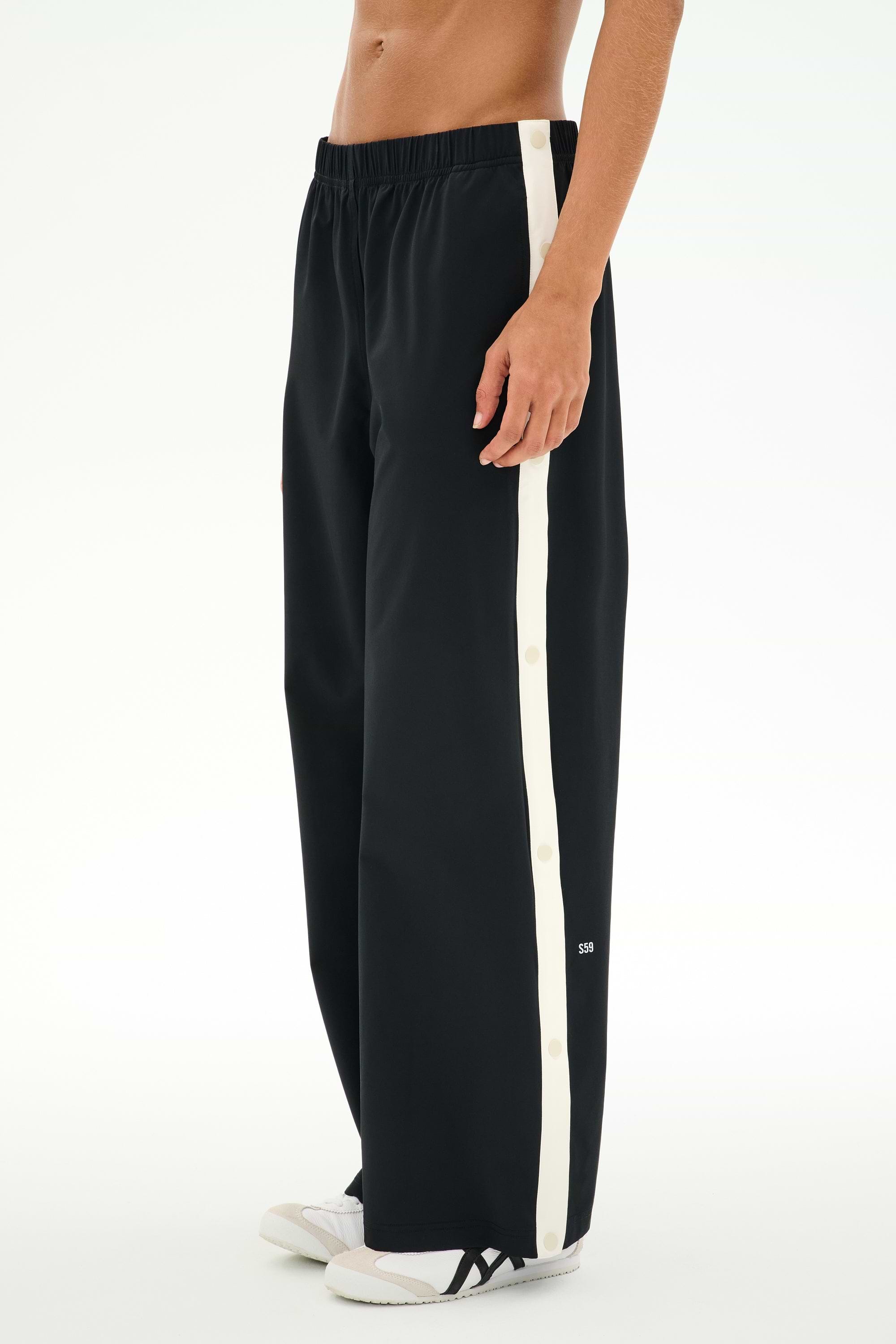 A person wears the SPLITS59 Jordan Nylon Twill Tearaway pants in Black/Creme, featuring a comfortable nylon-spandex blend, wide-leg fit, white side stripe, and snap buttons, paired with white sneakers.